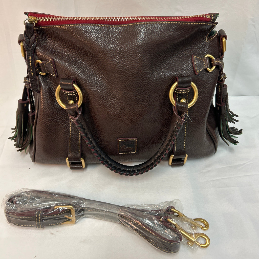 A dark brown pebbled leather shoulder bag with gold-tone hardware, featuring a top zipper, braided leather handles, and dark green tassel accents. A matching detachable shoulder strap is wrapped in protective plastic and placed in front of the bag on a white fabric background.