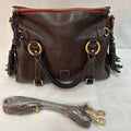 A dark brown pebbled leather shoulder bag with gold-tone hardware, featuring a top zipper, braided leather handles, and dark green tassel accents. A matching detachable shoulder strap is wrapped in protective plastic and placed in front of the bag on a white fabric background.