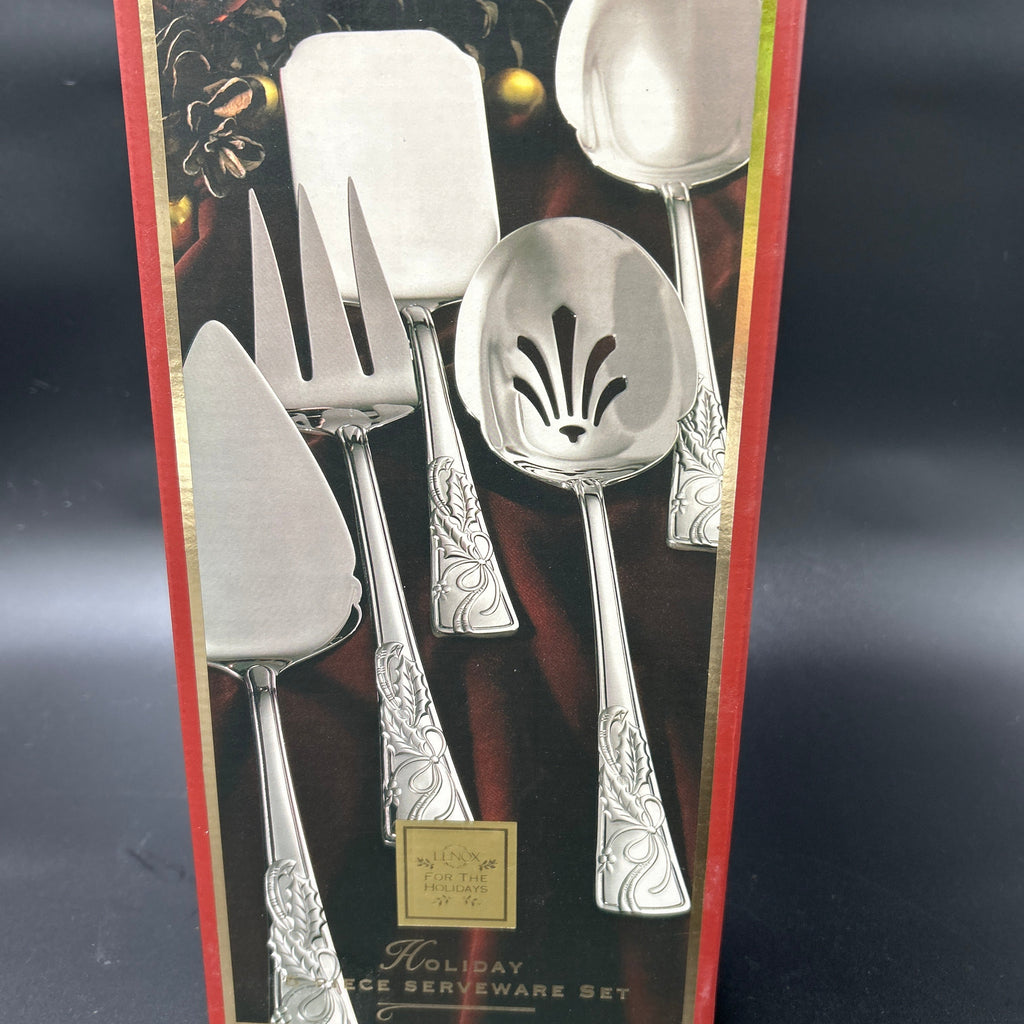 A boxed holiday serving ware set with silver-toned utensils visible through a red and gold package, photographed against a dark studio background. New Lenox Holiday 5-Piece Stainless Steel Serveware Hostess Set 