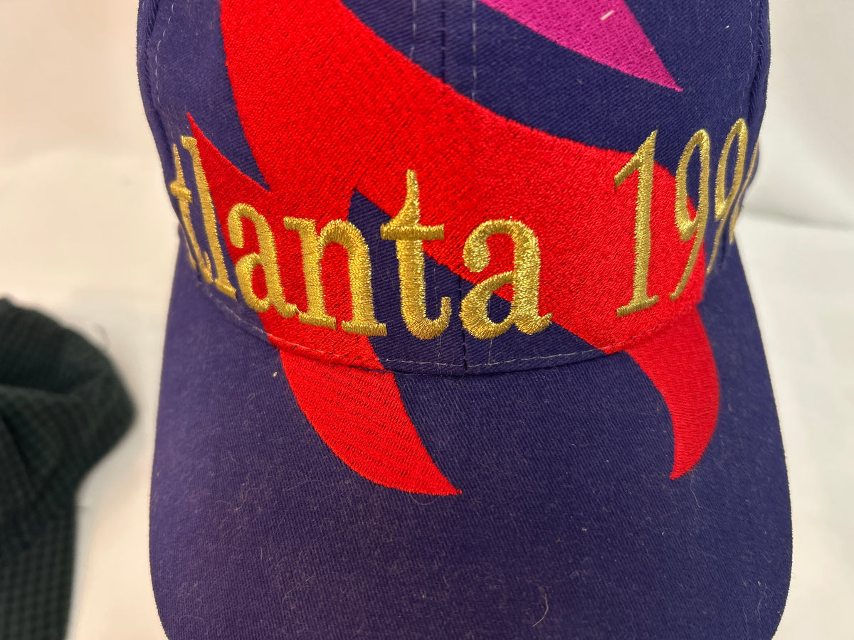 A close-up of a vintage-style baseball cap featuring a dark purple crown with a large red abstract graphic, gold embroidered text reading 'Atlanta 199', and a small pink accent at the top. A black textured fabric item is partially visible at the left edge.