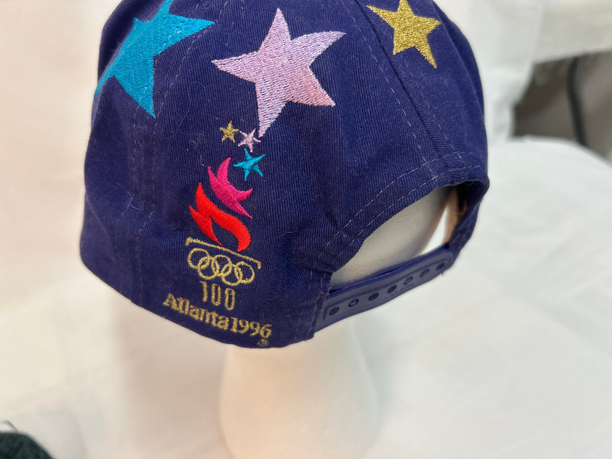 A navy blue baseball cap featuring embroidered stars in light blue, pink, and gold, along with a red and gold Atlanta 1996 Olympic flame and rings design. The cap is displayed on a white mannequin head against a white fabric background.