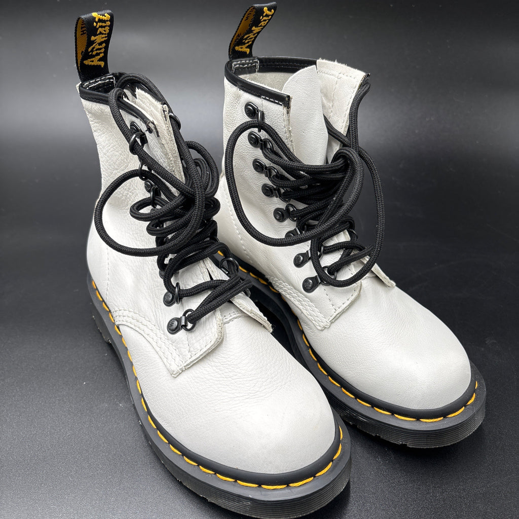 A pair of white leather Dr. Martens 16mm lace-up boots with black laces, black soles, and signature yellow stitching, displayed on a dark gray background.