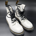 A pair of white leather Dr. Martens 16mm lace-up boots with black laces, black soles, and signature yellow stitching, displayed on a dark gray background.