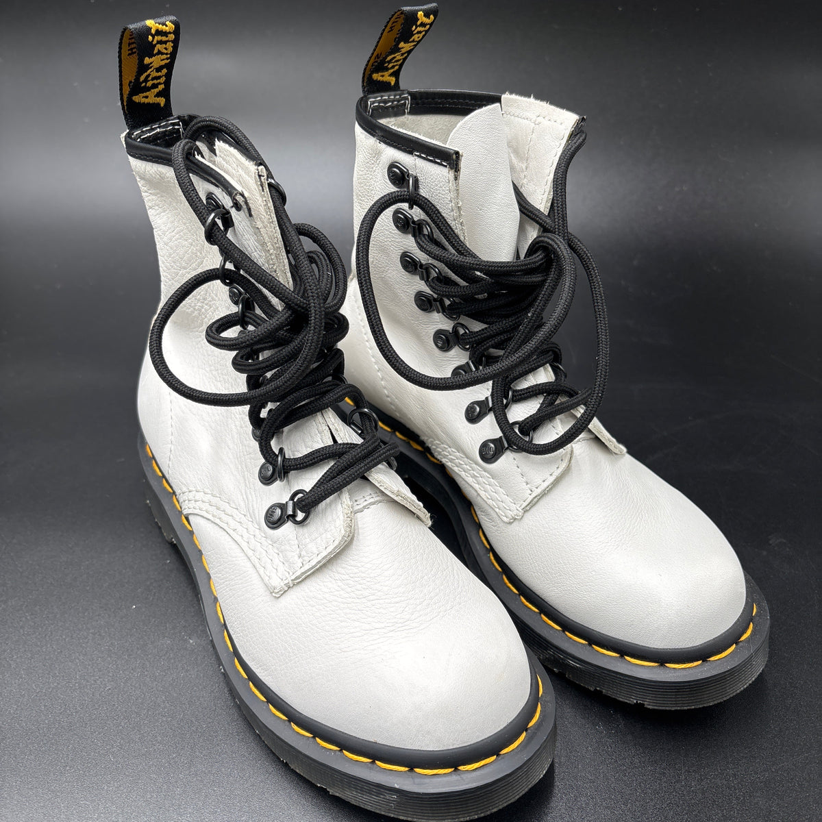 A pair of white leather Dr. Martens 16mm lace-up boots with black laces, black soles, and signature yellow stitching, displayed on a dark gray background.