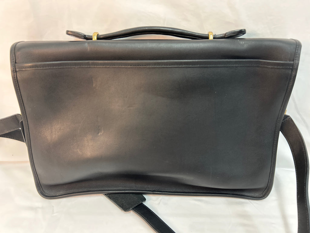 A black leather Coach shoulder bag with a top handle and a long strap, photographed against a white fabric background.