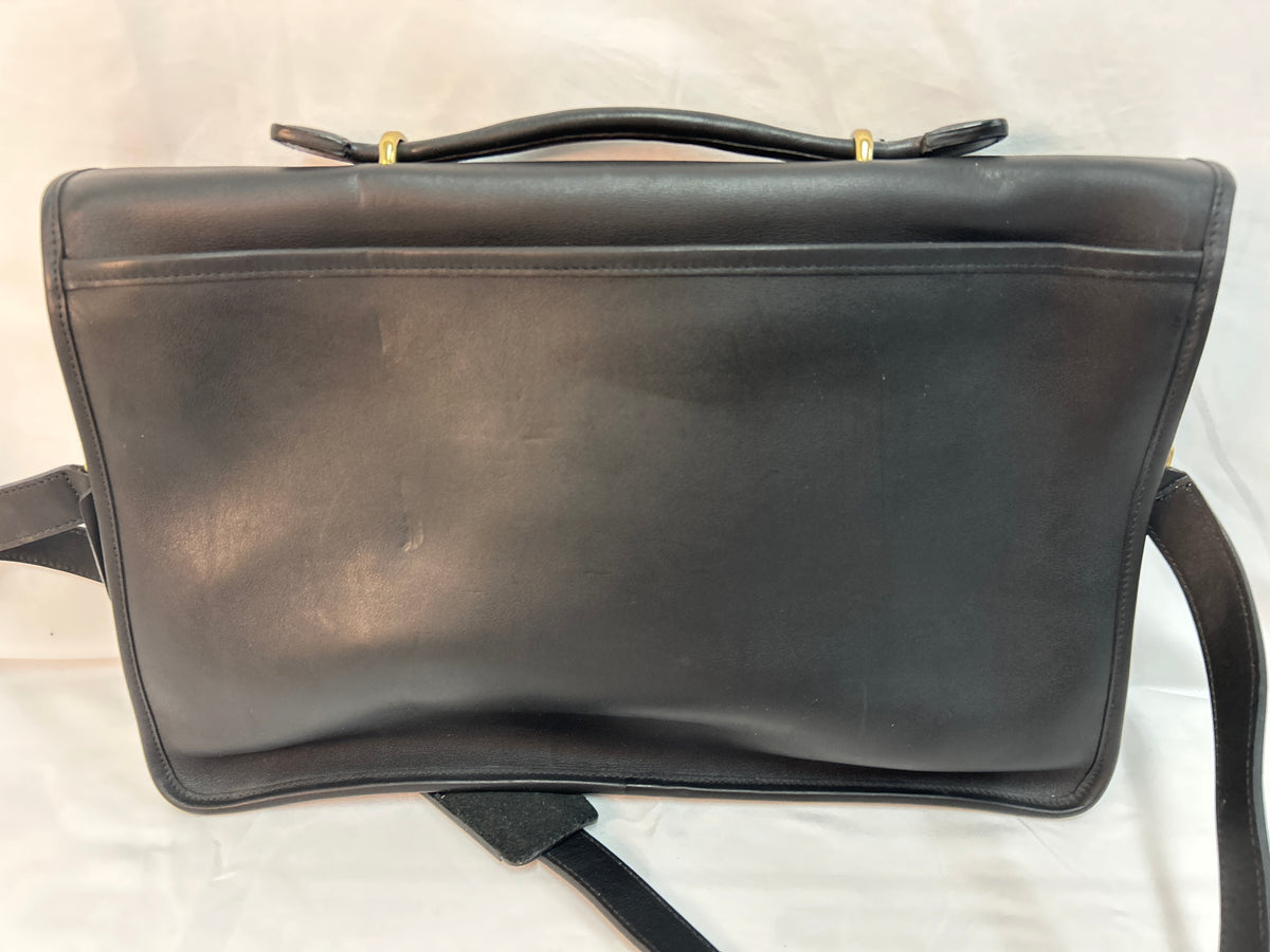 A black leather Coach shoulder bag with a top handle and a long strap, photographed against a white fabric background.