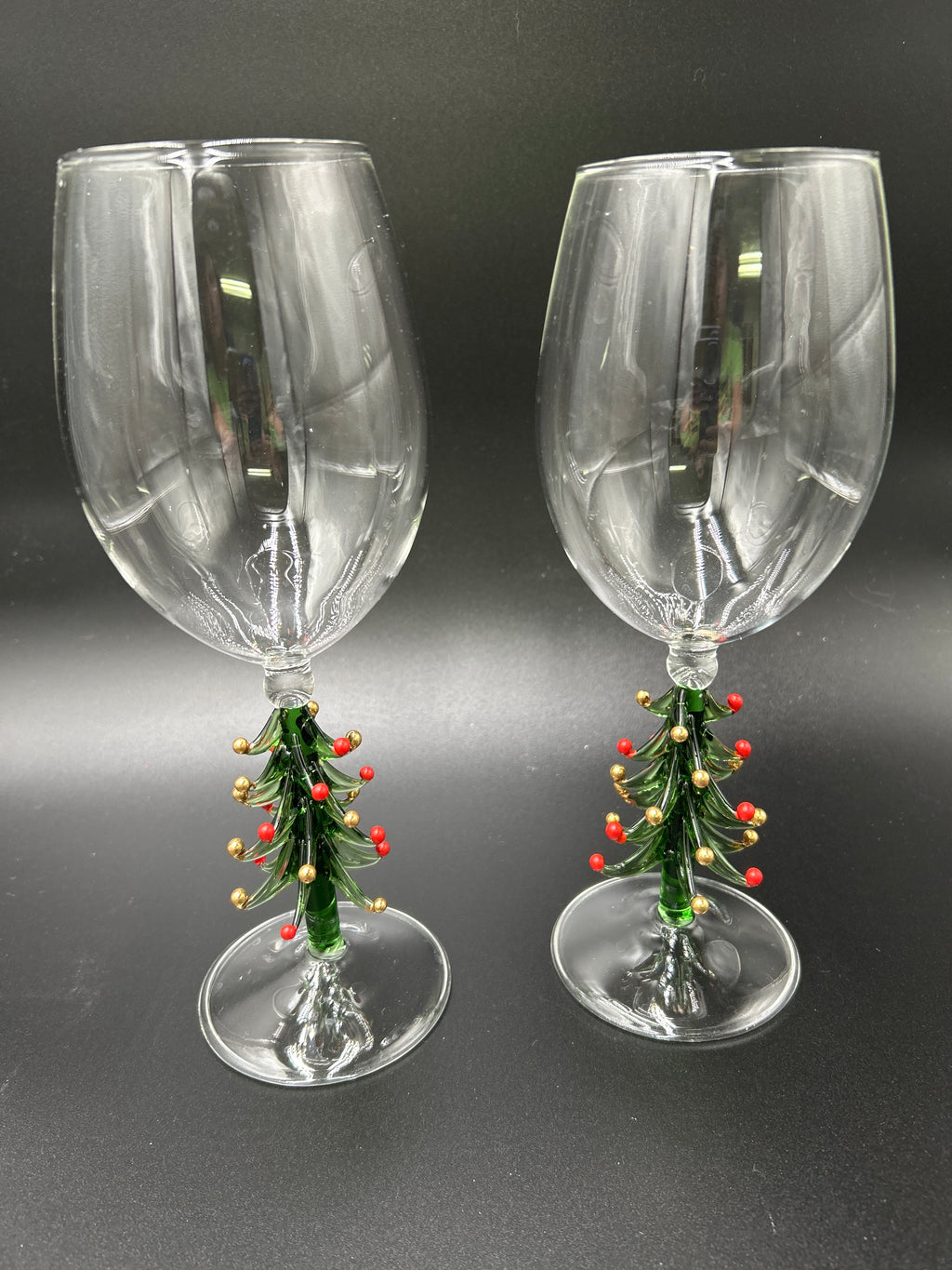 A pair of clear glass wine glasses with miniature green Christmas tree stems decorated with red berries and gold ornaments, set against a dark gray background. Neiman Marcus Figural Christmas Tree Wine Glasses 