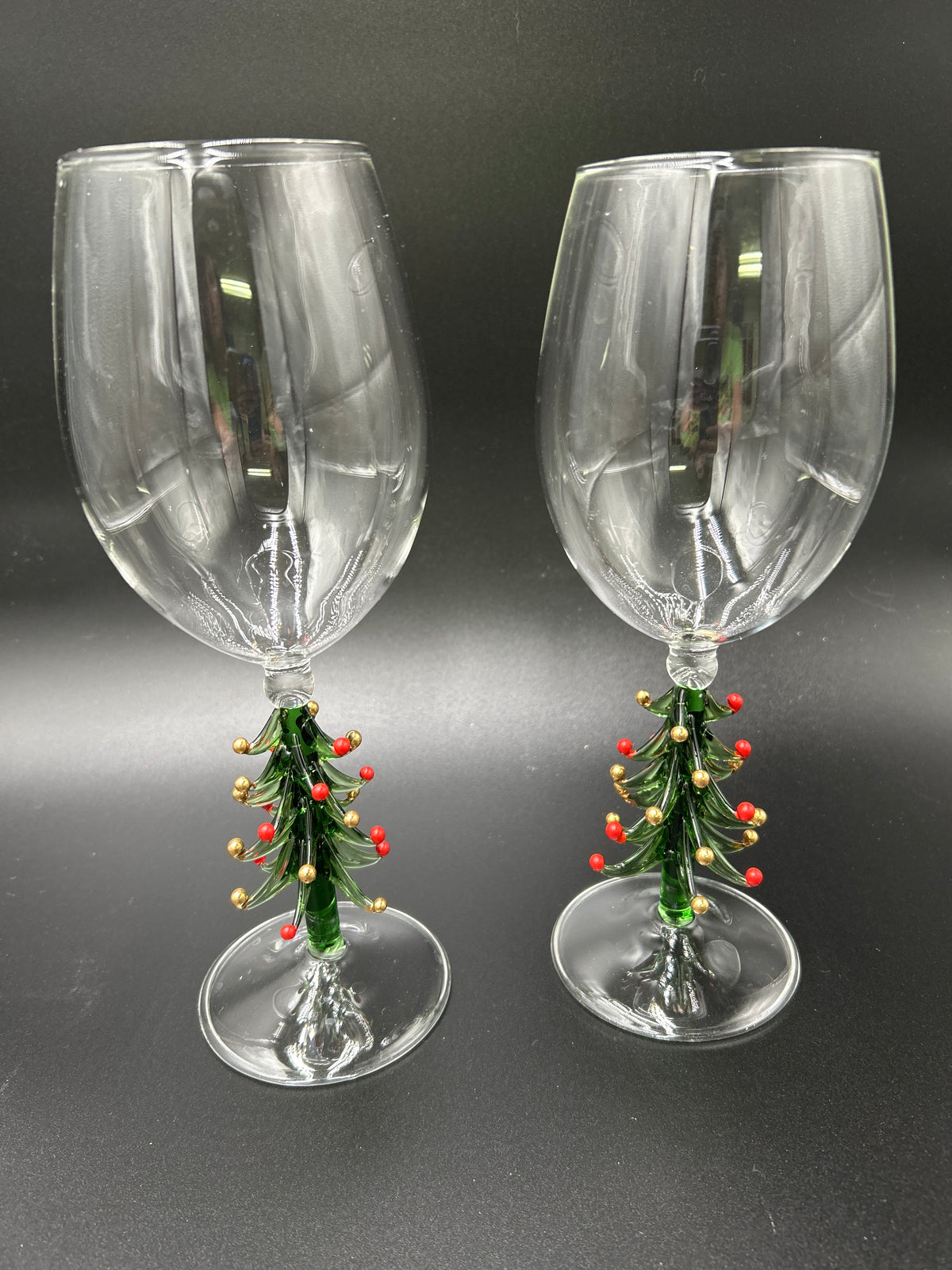 A pair of clear glass wine glasses with miniature green Christmas tree stems decorated with red berries and gold ornaments, set against a dark gray background. Neiman Marcus Figural Christmas Tree Wine Glasses 