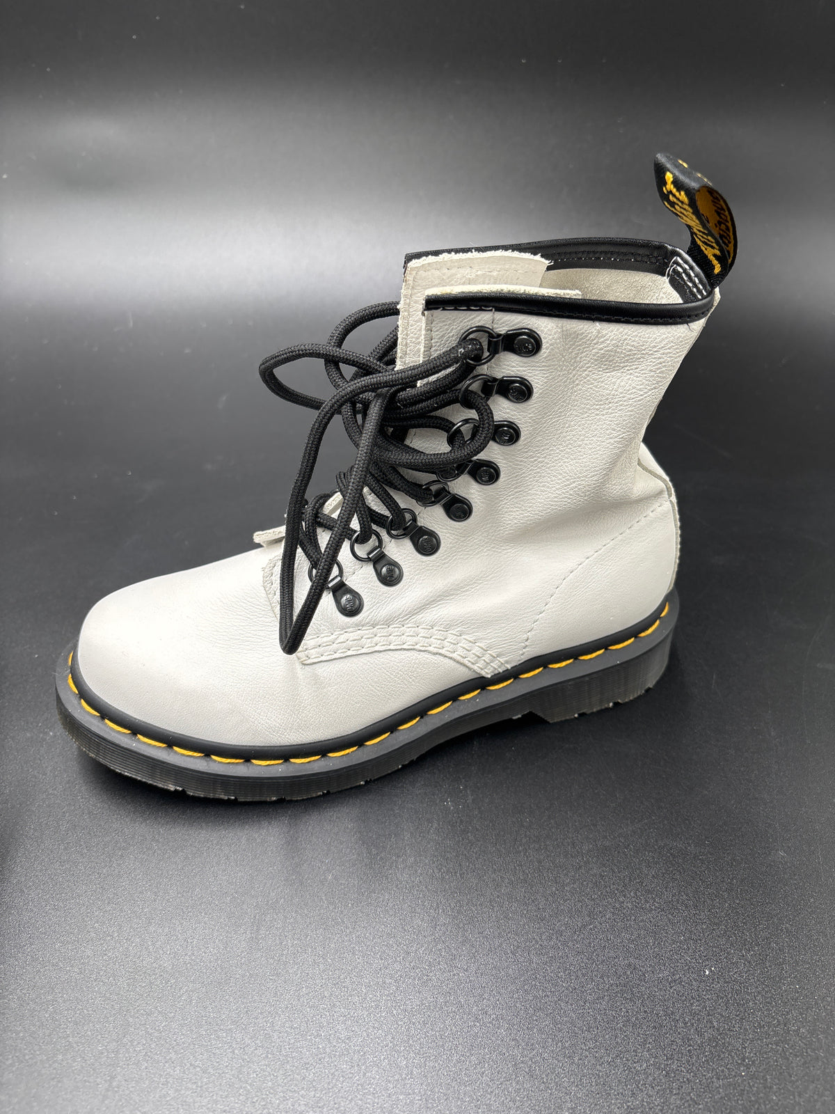 A single white leather Dr. Martens lace-up boot with black laces, black sole, and signature yellow stitching, shown on a dark gray background.