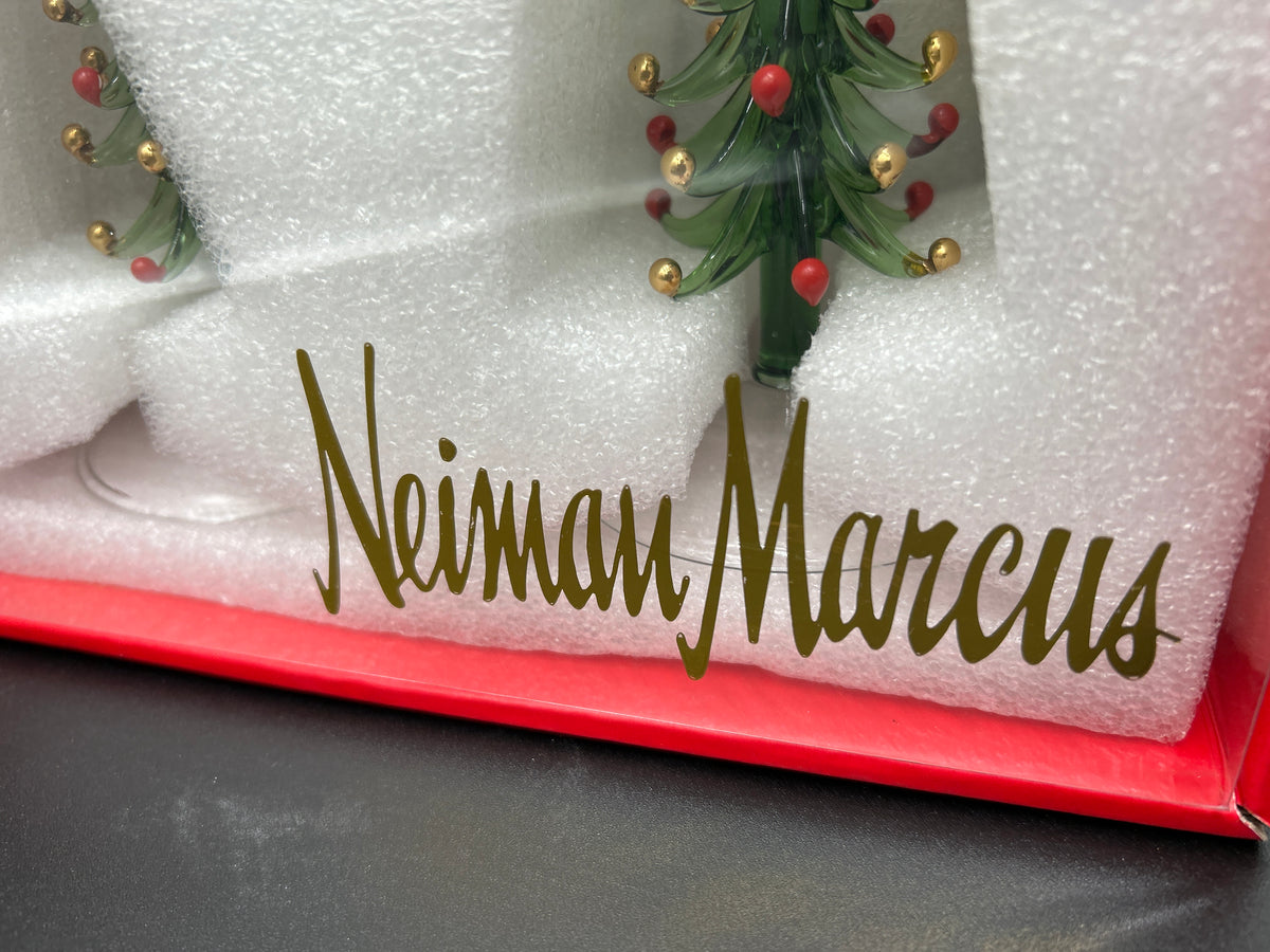 A close-up of a personalized glass Christmas tree ornament inside its red gift box, nestled in white foam. The ornament features the custom name 'NeimanMarcus' in gold cursive script, with a green glass tree decorated with red and gold ornaments. Neiman Marcus Figural Christmas Tree Wine Glasses 