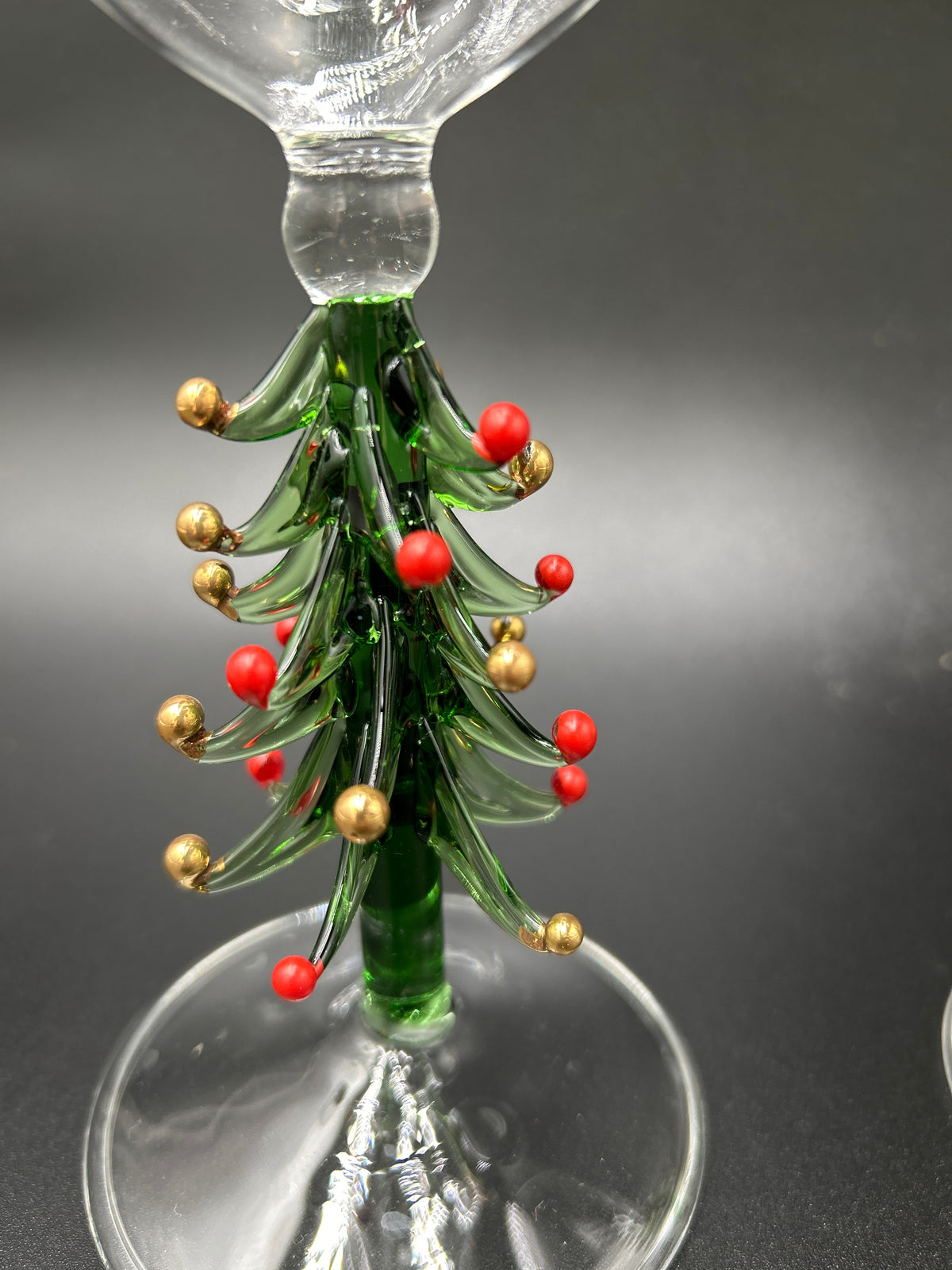 A close-up of a clear glass stemware piece featuring a decorative green glass Christmas tree with red berries and gold ornaments on a dark gray background. Neiman Marcus Figural Christmas Tree Wine Glasses 