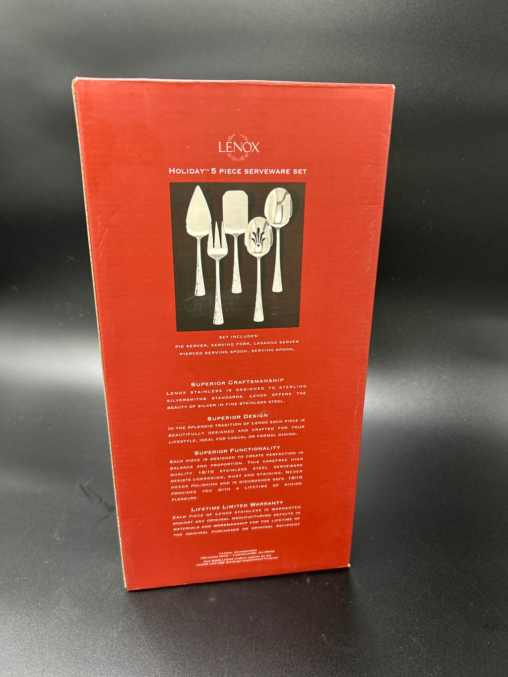 A tall red Lenox product box for a holiday 5-piece serverware set, standing upright against a dark gray background. The front of the box features a black square with silver-colored cutlery illustrations and white text. New Lenox Holiday 5-Piece Stainless Steel Serveware Hostess Set 