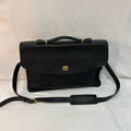 A black leather briefcase-style Coach handbag with a top handle, a detachable shoulder strap, and a small gold-tone logo clasp, photographed against a white fabric backdrop.