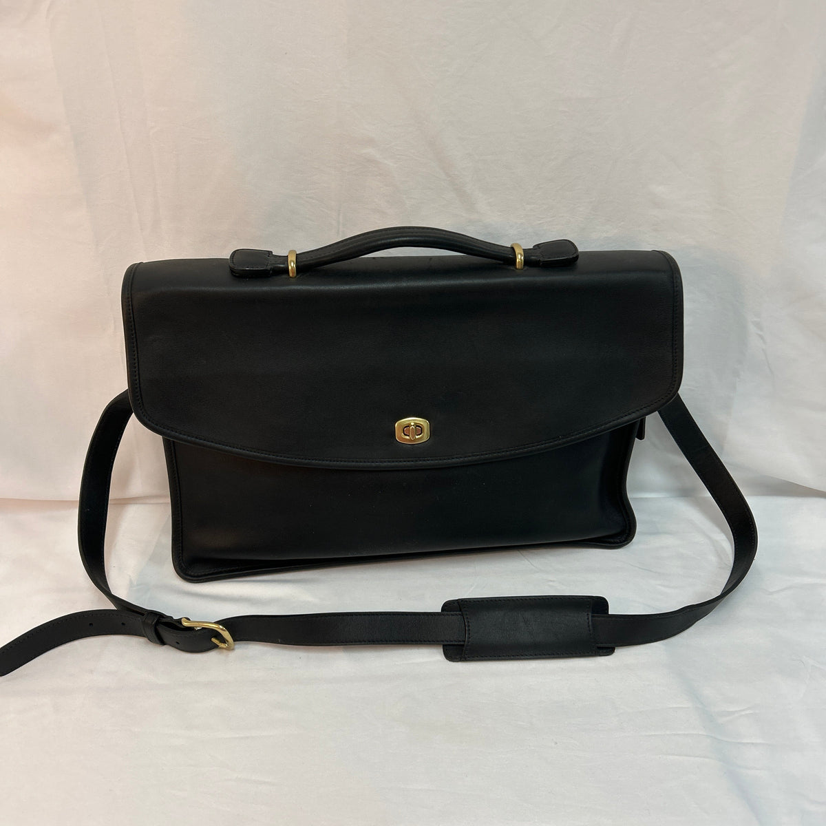 A black leather briefcase-style Coach handbag with a top handle, a detachable shoulder strap, and a small gold-tone logo clasp, photographed against a white fabric backdrop.