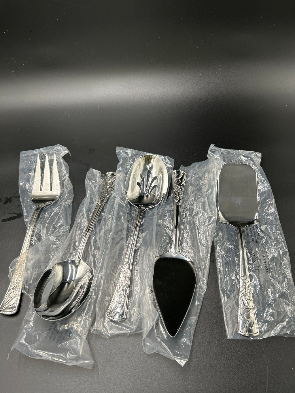 A set of five stainless steel tableware utensils, including a dinner fork, dinner spoon, soup spoon, pie server, and serving spatula, all wrapped in clear plastic protective packaging on a dark gray background. New Lenox Holiday 5-Piece Stainless Steel Serveware Hostess Set 