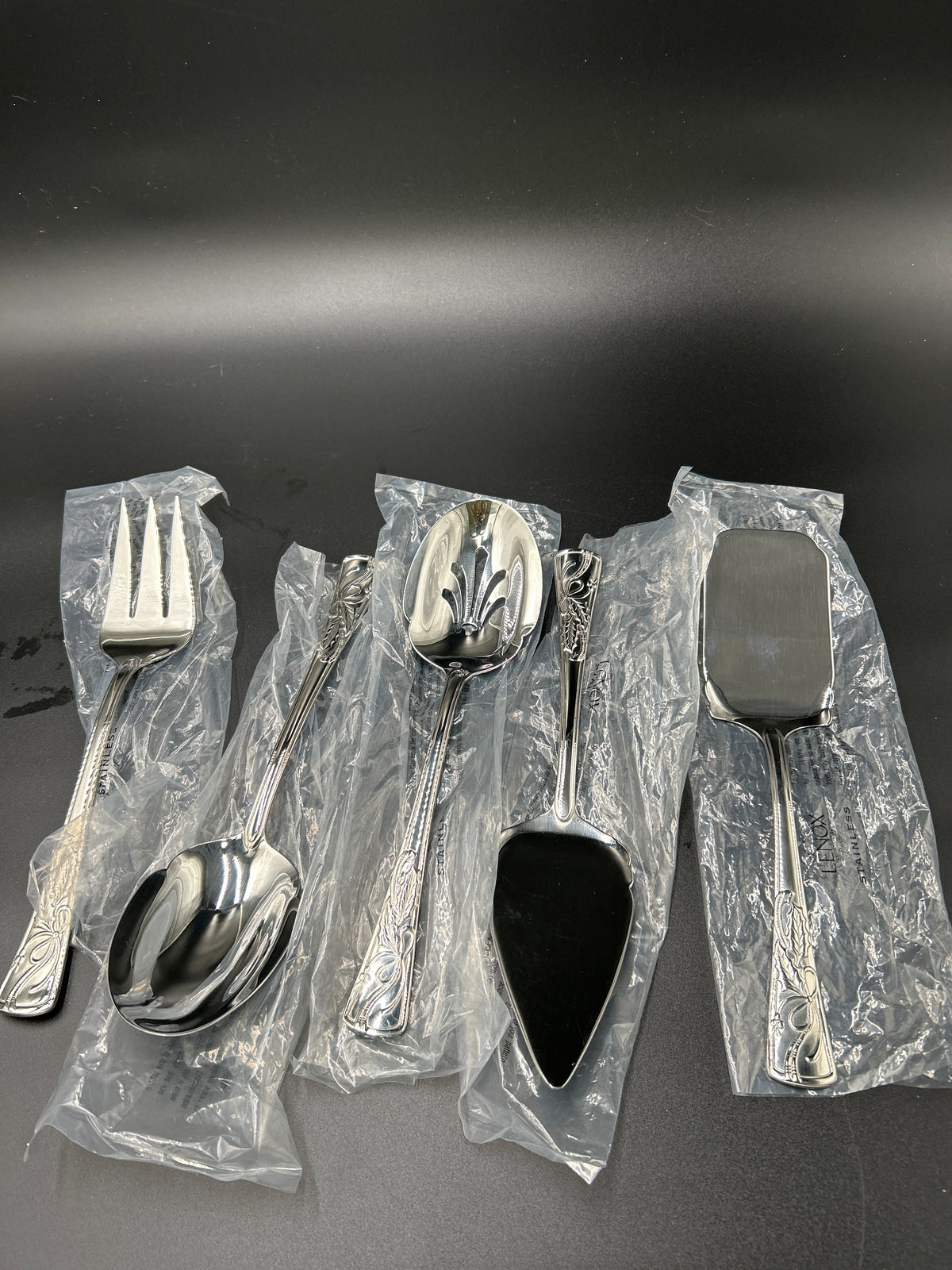 A set of five stainless steel tableware utensils, including a dinner fork, dinner spoon, soup spoon, pie server, and serving spatula, all wrapped in clear plastic protective packaging on a dark gray background. New Lenox Holiday 5-Piece Stainless Steel Serveware Hostess Set 