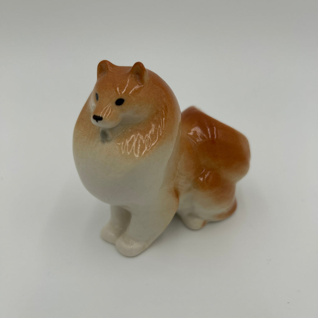 A small ceramic figurine of a fluffy dog, likely a Shiba Inu, with a glossy finish. The dog features a warm tan and reddish-brown coat with a cream-colored underbelly and paws, standing on a seamless light gray background.