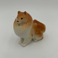 A small ceramic figurine of a fluffy dog, likely a Shiba Inu, with a glossy finish. The dog features a warm tan and reddish-brown coat with a cream-colored underbelly and paws, standing on a seamless light gray background.