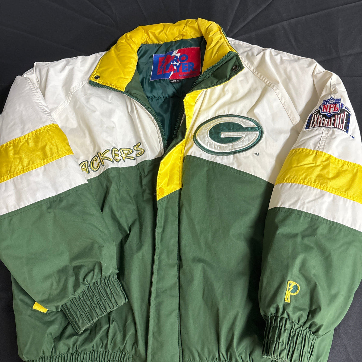 A vintage Green Bay Packers jacket from the 90s, featuring a white and green color-blocked design with yellow accents. The jacket has 'PACKERS' embroidered on the right chest, a large Packers 'G' logo on the left chest, and a 'NFL Antiques' patch on the right sleeve.