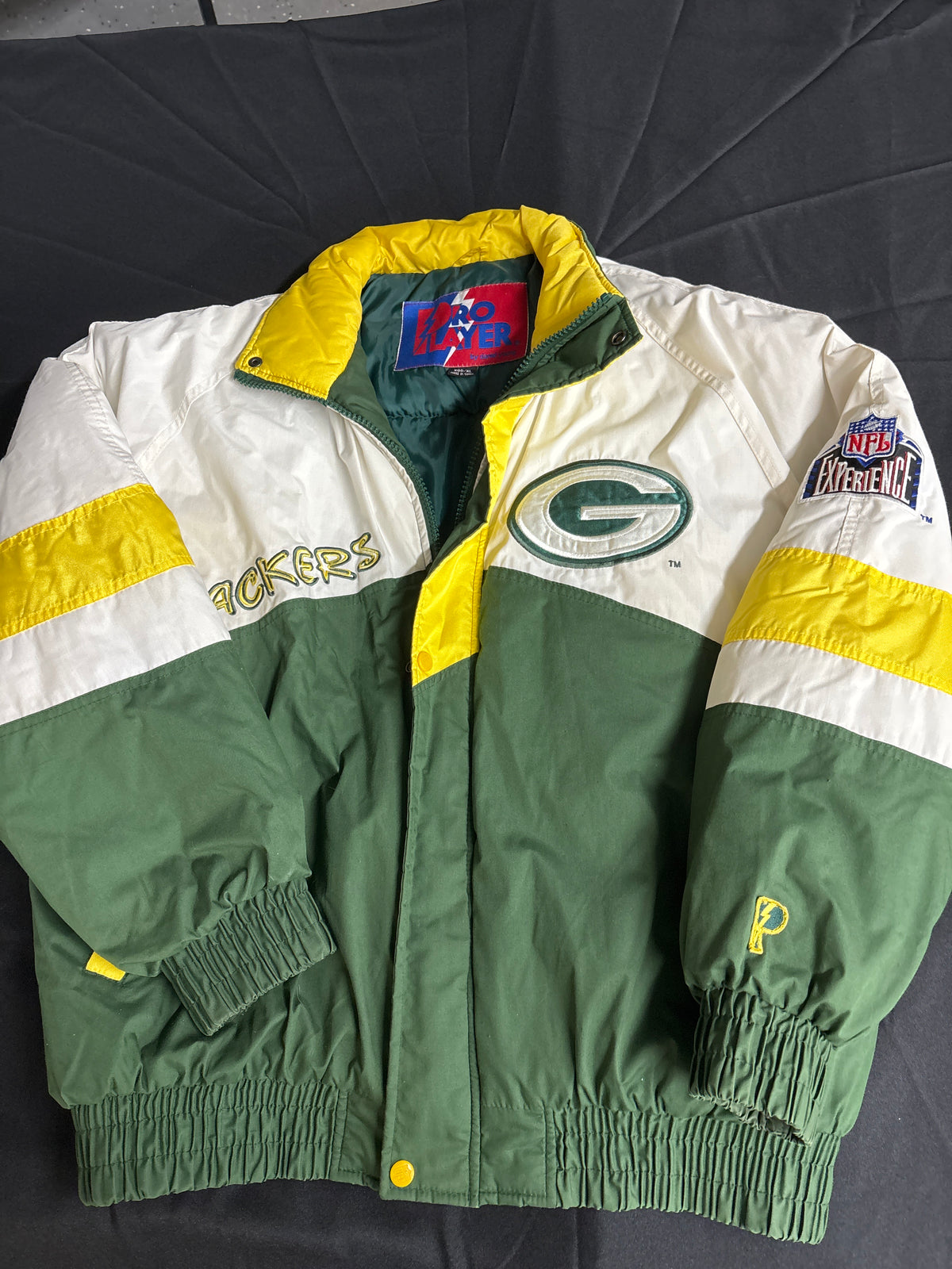 A vintage Green Bay Packers jacket from the 90s, featuring a white and green color-blocked design with yellow accents. The jacket has 'PACKERS' embroidered on the right chest, a large Packers 'G' logo on the left chest, and a 'NFL Antiques' patch on the right sleeve.