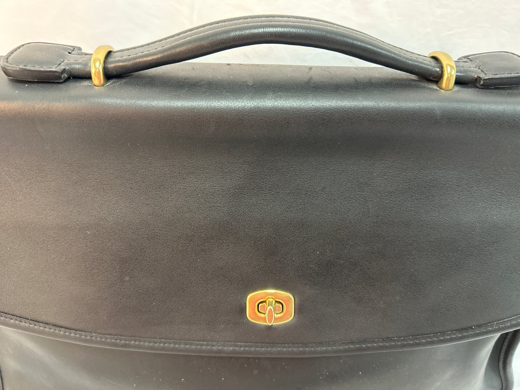 A close-up of a black leather briefcase or satchel with gold-tone hardware, featuring a top handle and a small oval logo plaque on the front flap.