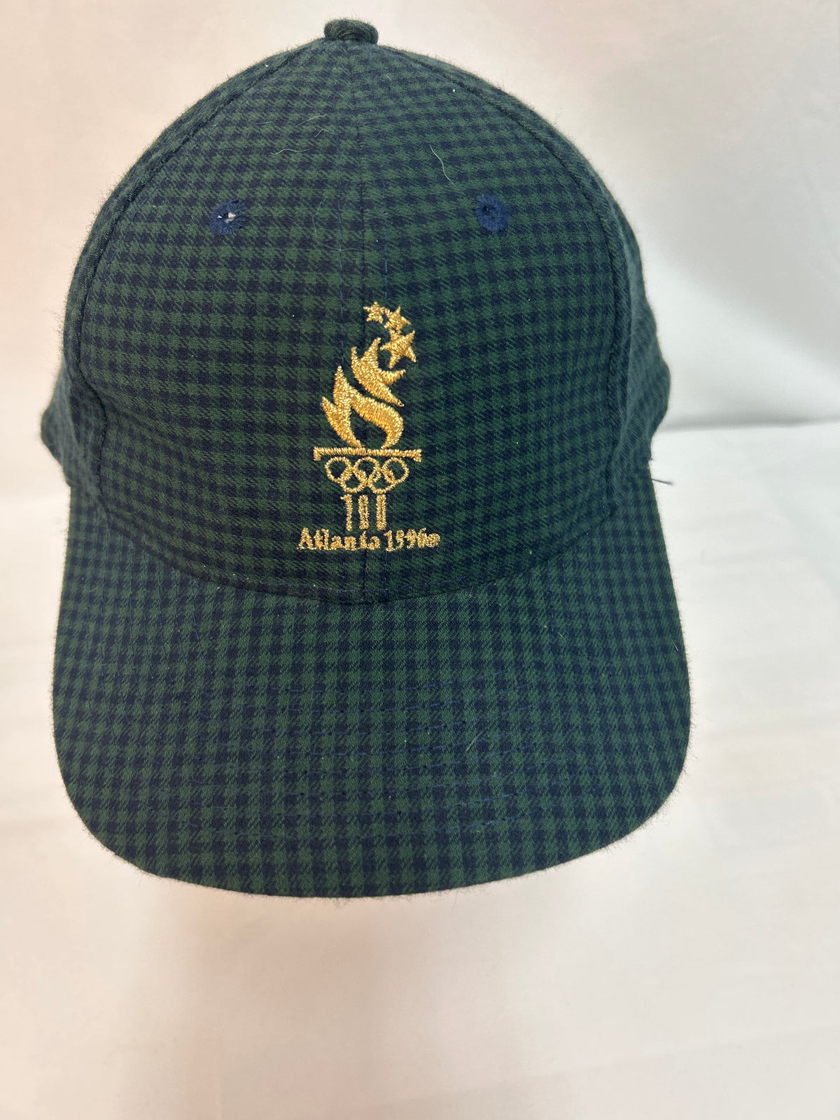 A dark green and black checkered baseball cap featuring a gold embroidered Atlanta 1996 Olympic logo on the front, displayed against a plain light background.