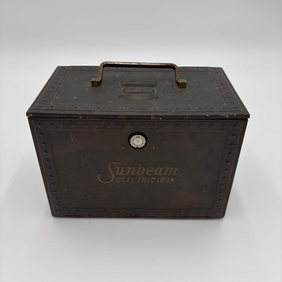 An antique dark brown metal Sunbeam Electric Iron case with a top handle, embossed rivets, and gold-toned branding on a light gray background.