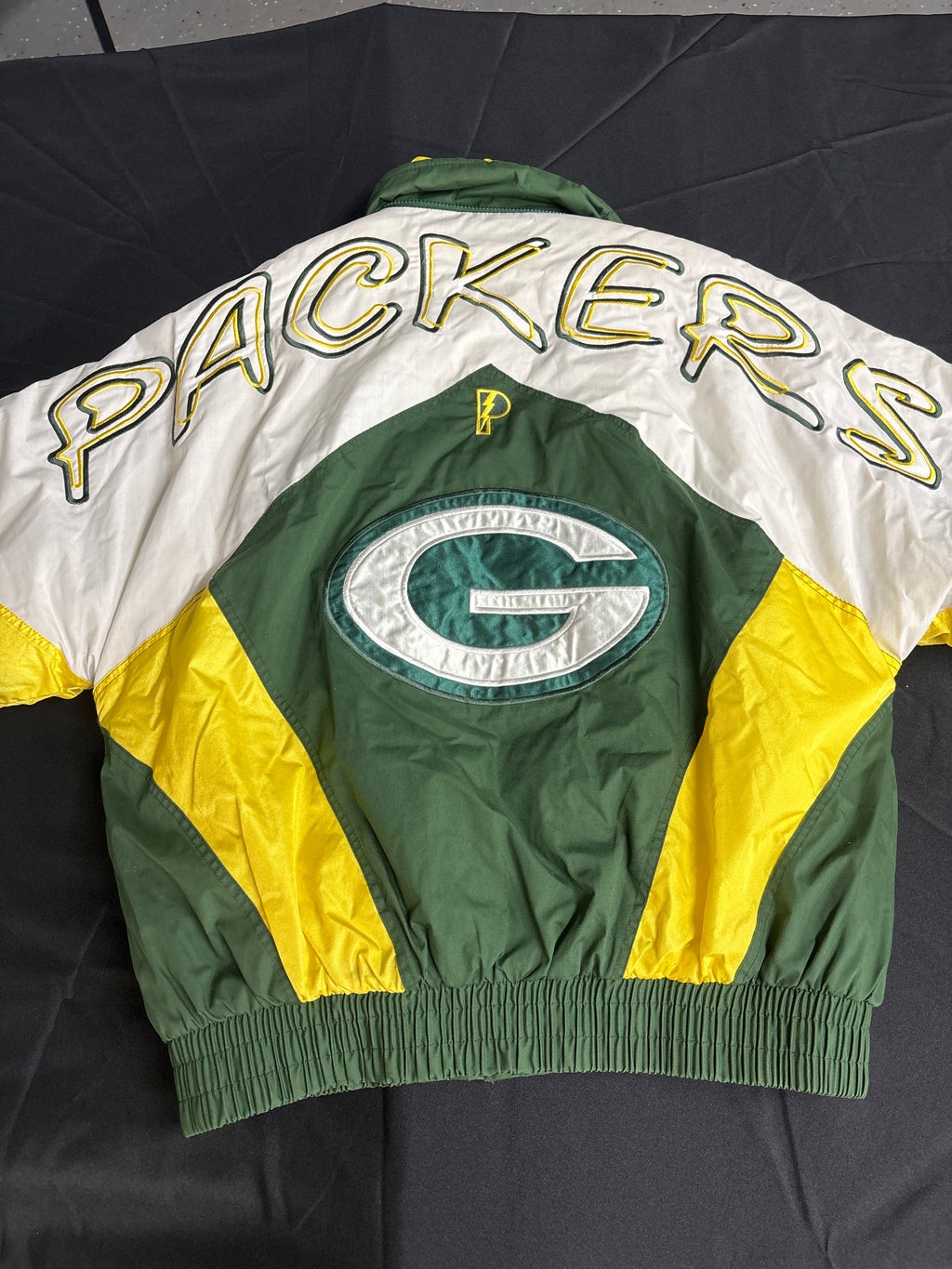 A vintage Green Bay Packers jacket laid flat on a black background, showing the back with 'PACKERS' in large yellow outlined letters across white shoulders, a green and white Packers 'G' logo patch in the center, and green and yellow panels.