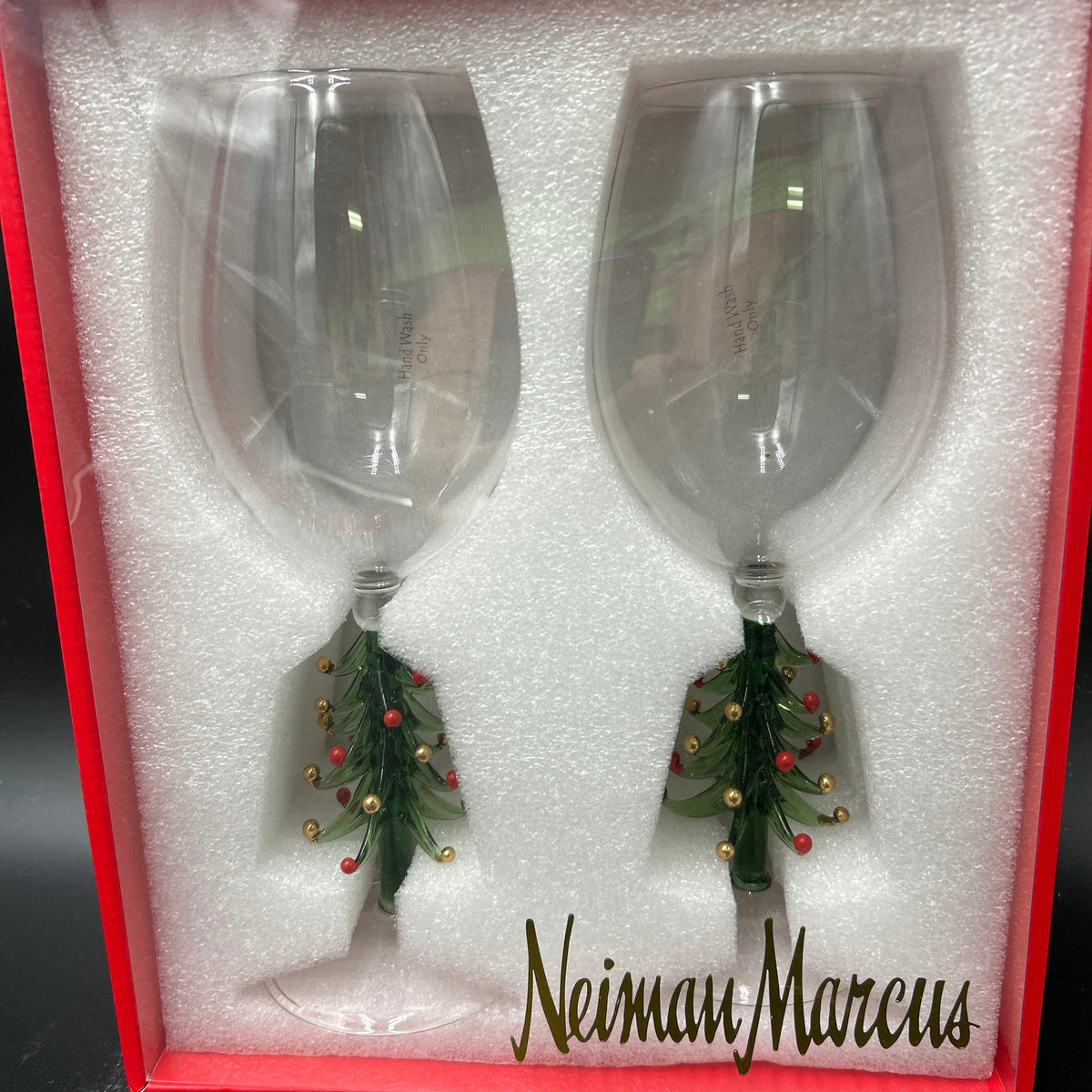 A pair of clear wine glasses with green Christmas tree-shaped stems decorated with red and gold ornaments, presented in a red gift box with white foam insert. The brand name 'Neiman Marcus' is printed on the front. Neiman Marcus Figural Christmas Tree Wine Glasses 
