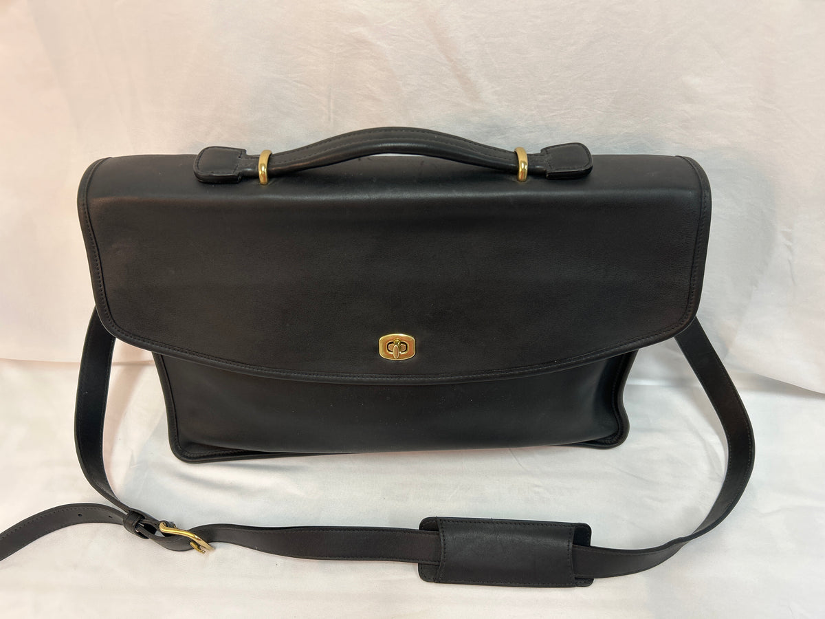 A black leather briefcase-style handbag with a top handle, detachable shoulder strap, and a small gold-tone oval logo plaque on the front, photographed against a white fabric backdrop.