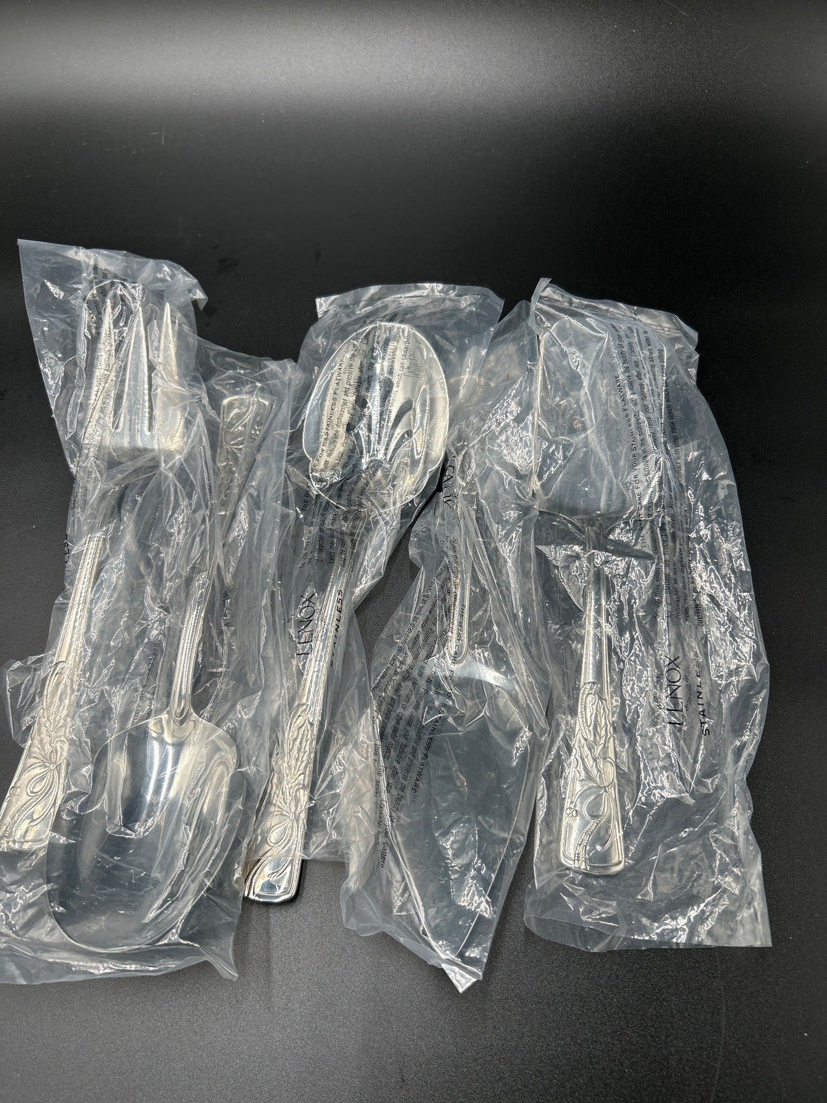 New Lenox Holiday 5-Piece Stainless Steel Serveware Hostess Set  Four packaged silver-toned serving utensils, including a soup ladle, spoon, and forks, sealed in clear plastic bags on a dark background. 