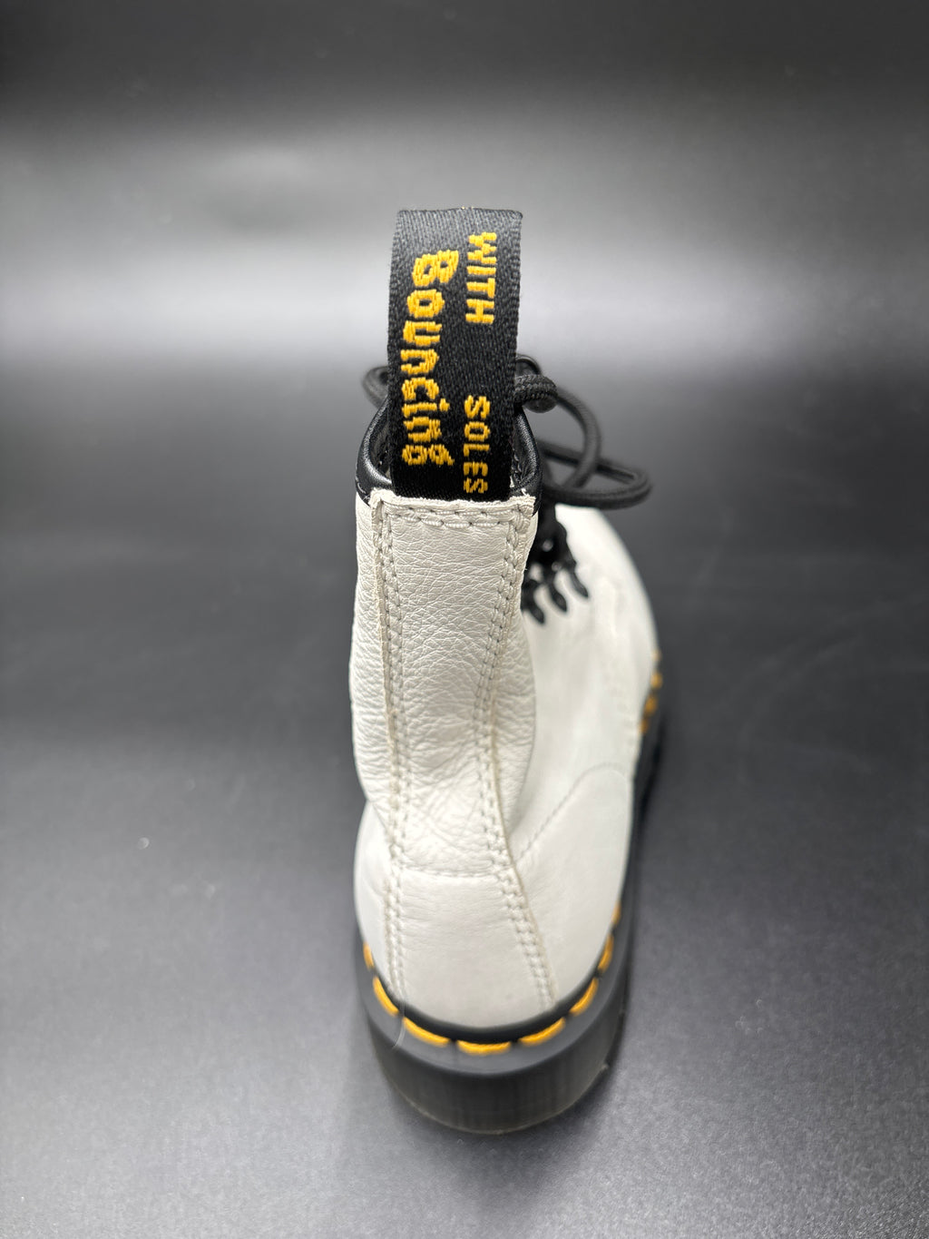 A close-up rear view of a white leather Dr. Martens boot with black laces and a black sole, showing the yellow 'Bourling' heel pull tab and contrast yellow stitching.