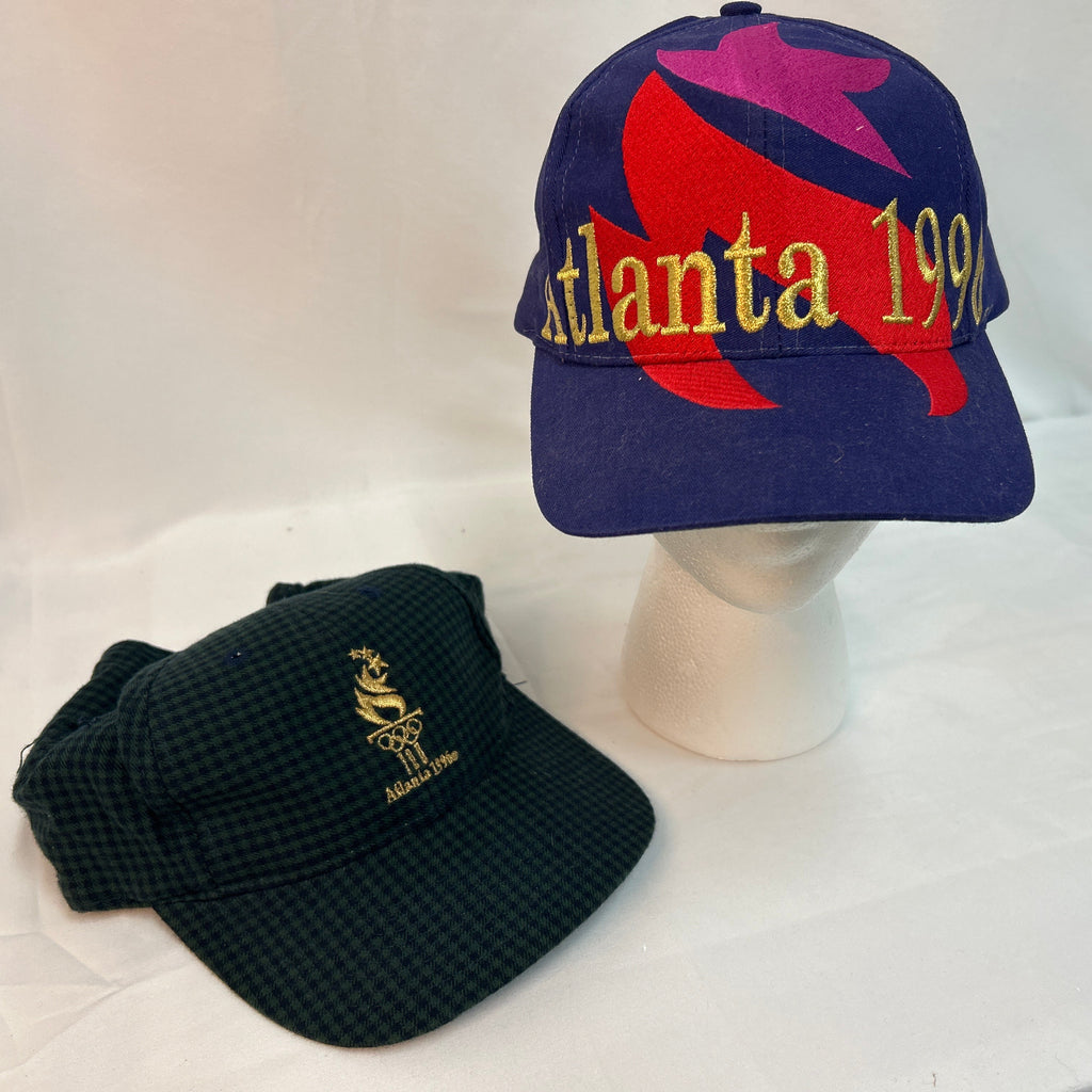 Two vintage Atlanta baseball caps displayed on a white fabric backdrop. The larger cap, sitting on a white mannequin head, is navy blue with a red and purple dolphin graphic and yellow 'Atlanta 1999' embroidery.