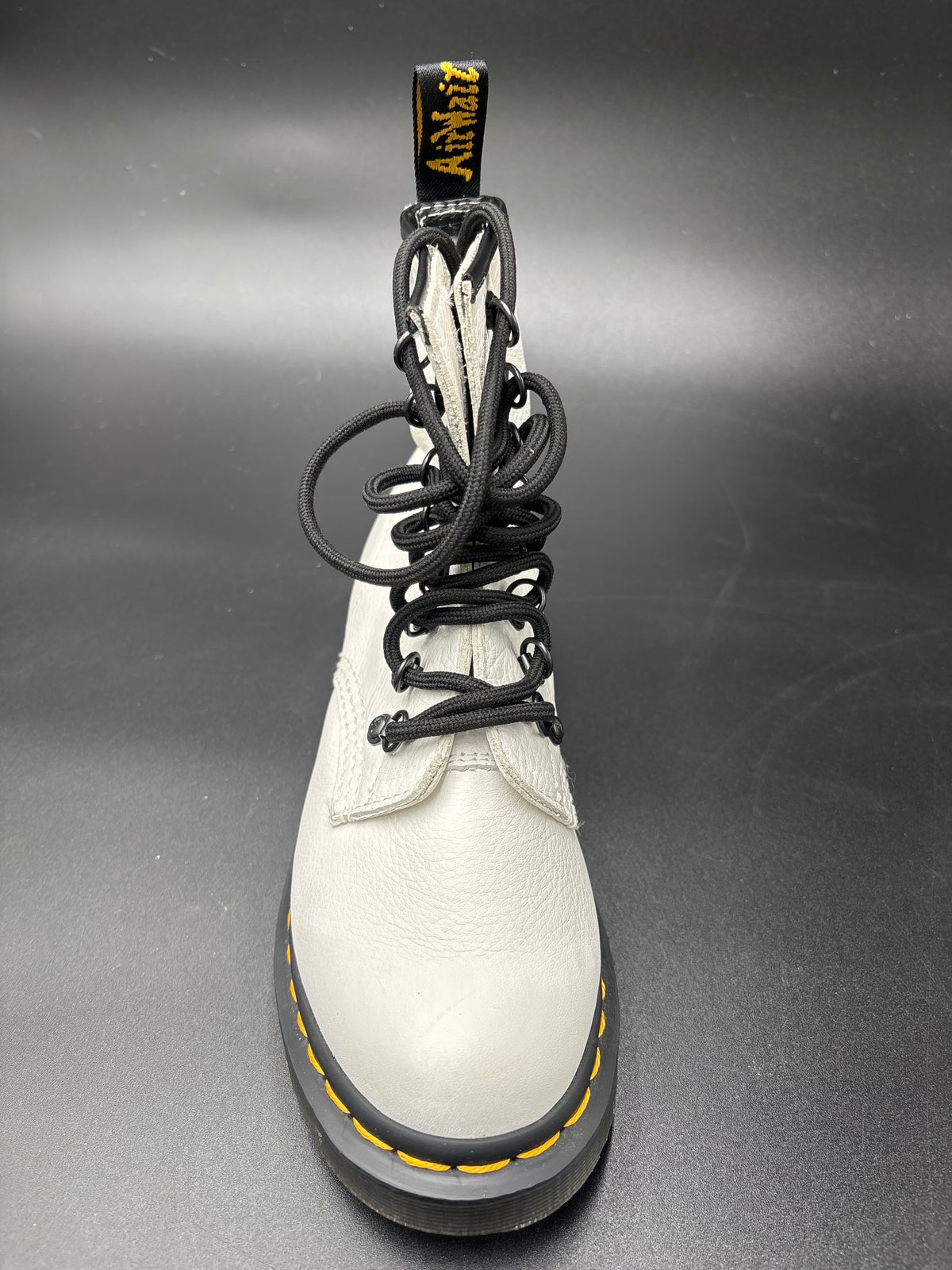 A single white leather Dr. Martens boot with black laces, a black sole, and yellow contrast stitching, photographed from the front on a dark gray background.