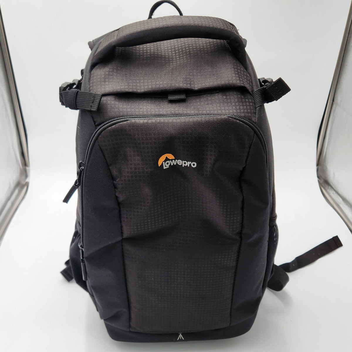 A black Lowepro backpack photographed upright against a white studio backdrop with clear light boxes visible on both sides. The backpack has a textured fabric body, multiple zip compartments, and the Lowepro logo on the front pocket.