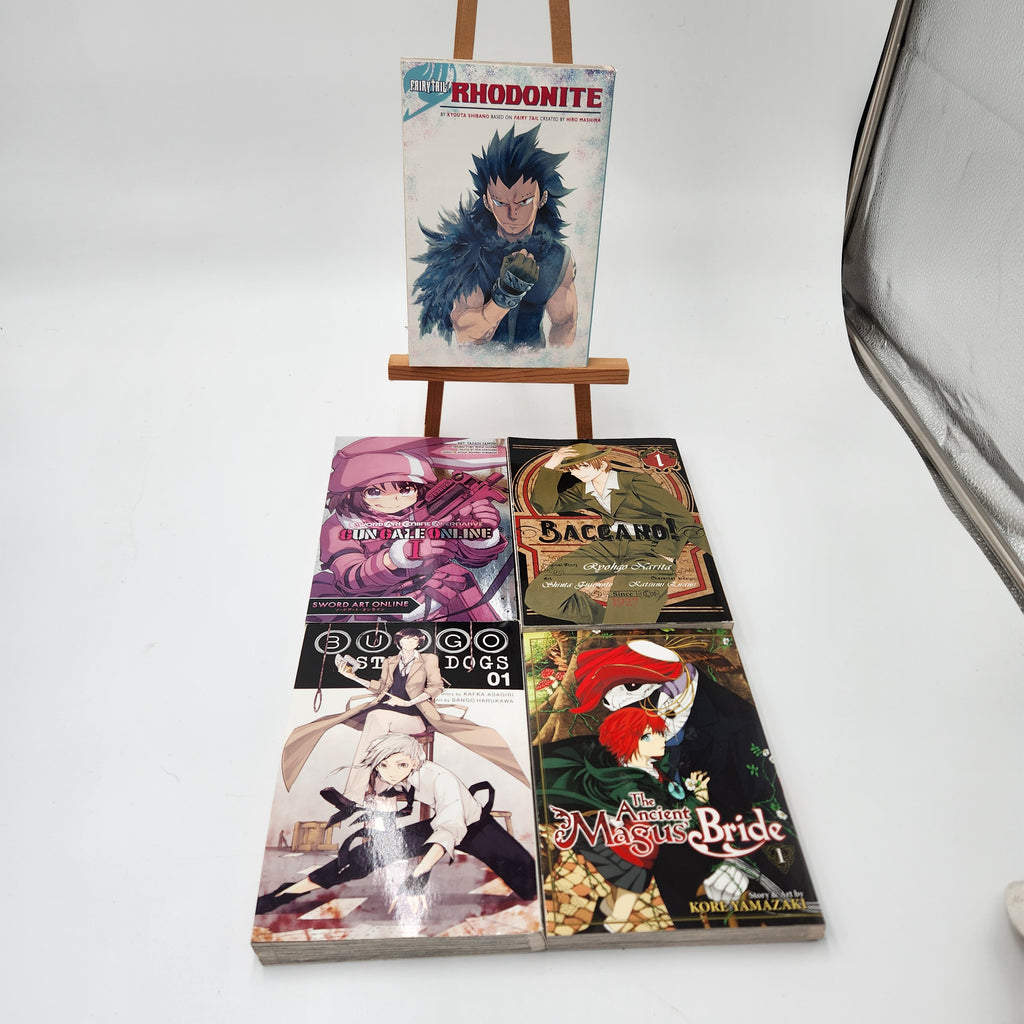 Five manga volumes from the franchise Rhodonite are displayed on a small wooden easel and the floor against a white studio background. The top volume stands on the easel, while the other three are laid out below.