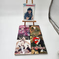 Five manga volumes from the franchise Rhodonite are displayed on a small wooden easel and the floor against a white studio background. The top volume stands on the easel, while the other three are laid out below.