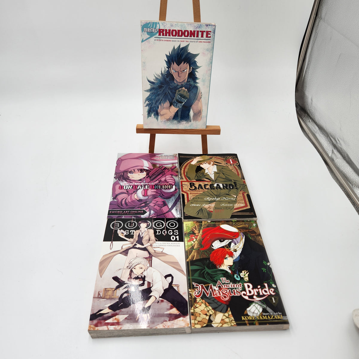 Five manga volumes from the franchise Rhodonite are displayed on a small wooden easel and the floor against a white studio background. The top volume stands on the easel, while the other three are laid out below.