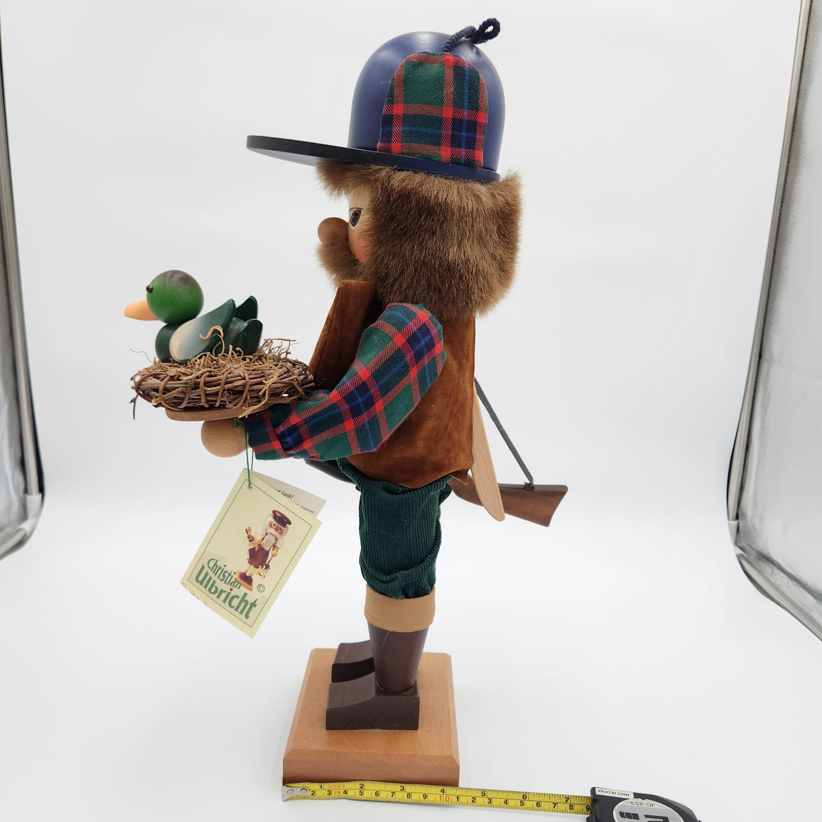 A traditional German wooden  Christian Ulbricht Nutcracker The Duck Hunter wearing a plaid hat and coat, holding a nest with two ducks. The figurine stands on a wooden base with a measuring tape laid out at the bottom, set against a white studio background with softbox lights visible on the sides.