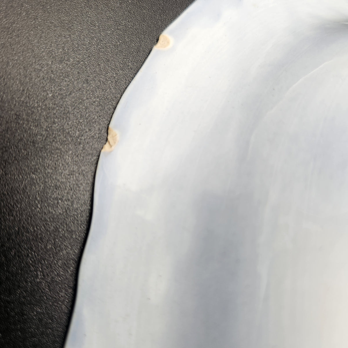 A close-up of a glossy white curved surface, likely a tray, bowl, or decorative piece, showing a few small beige wear marks against a dark textured background. Antique 1890s Flow Blue Cobalt Oval Serving Bowl Deep Vegetable Dish