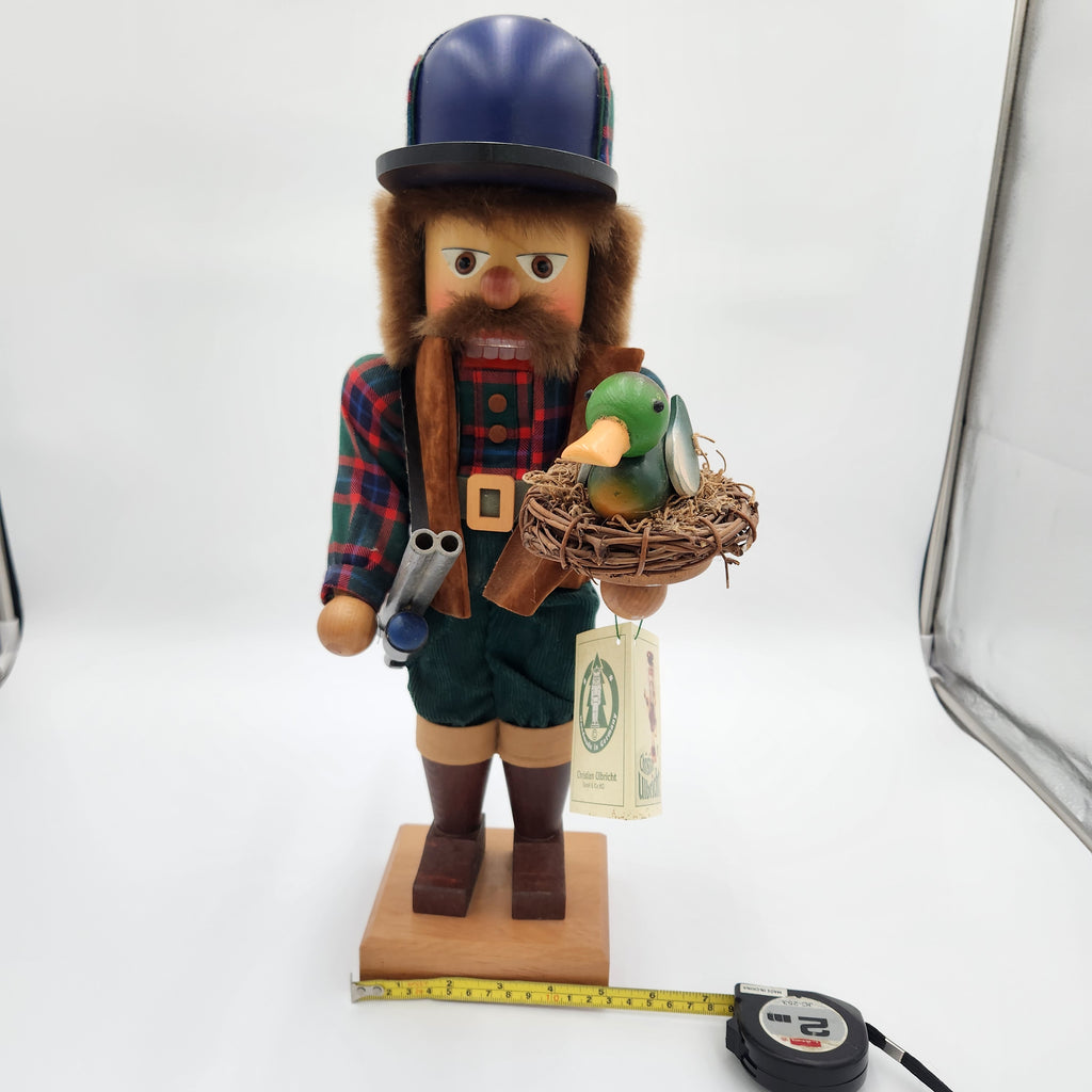 A handcrafted wooden   Christian Ulbricht Nutcracker The Duck Hunter:  a bearded man in a blue bowler hat and plaid shirt, holding a small green duck in a nest on a wooden base. A measuring tape is placed in front of the base on a white studio background.