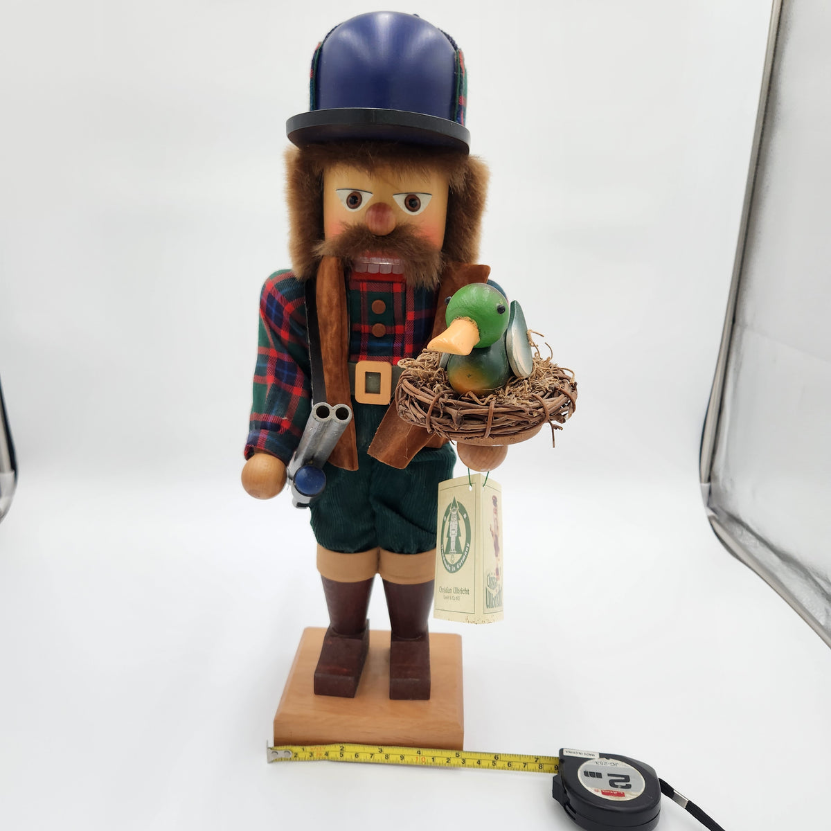A handcrafted wooden   Christian Ulbricht Nutcracker The Duck Hunter:  a bearded man in a blue bowler hat and plaid shirt, holding a small green duck in a nest on a wooden base. A measuring tape is placed in front of the base on a white studio background.
