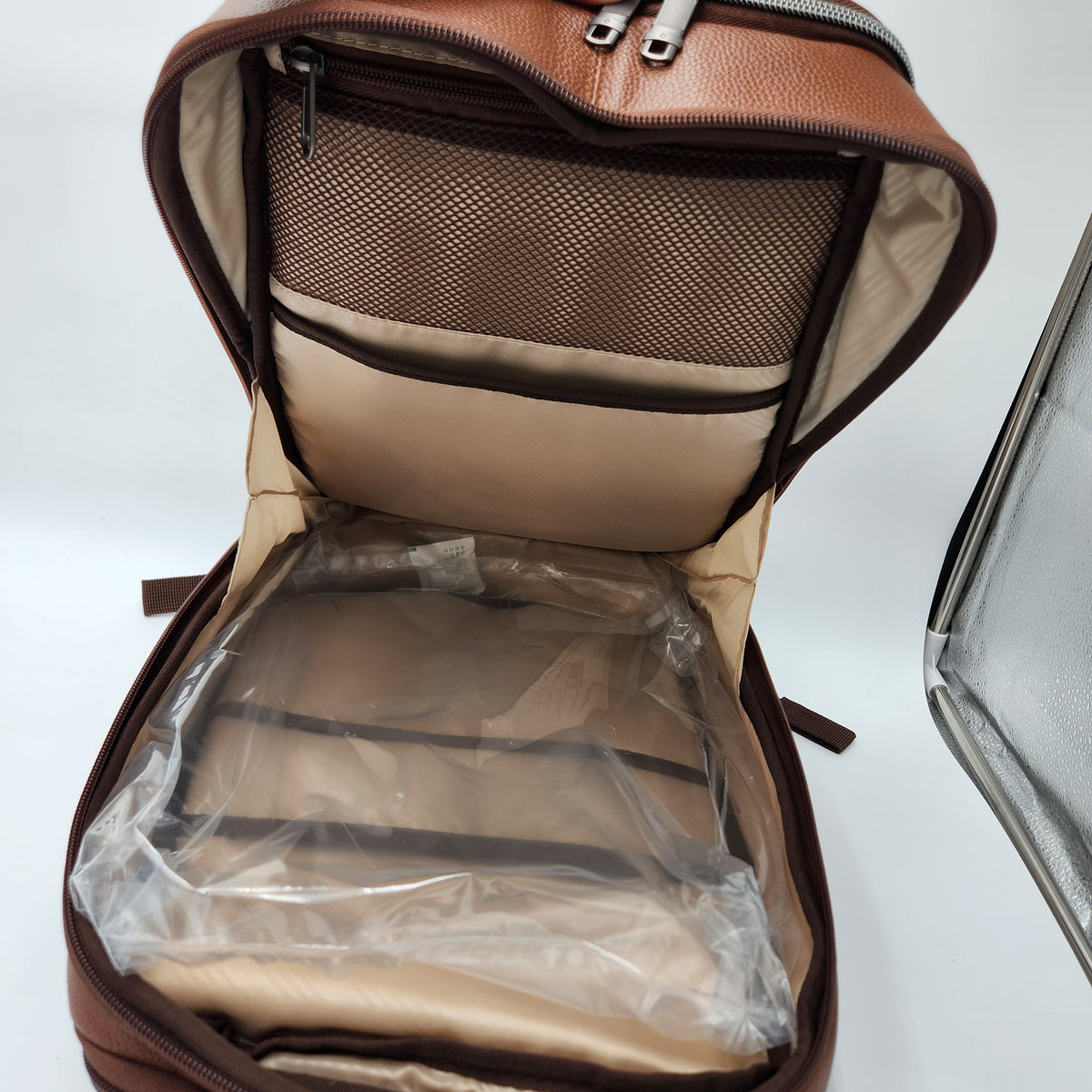 An open brown Samsonite leather backpack or laptop bag showing the interior with beige lining, a mesh pocket, and a clear plastic protective cover over the lower compartment.
