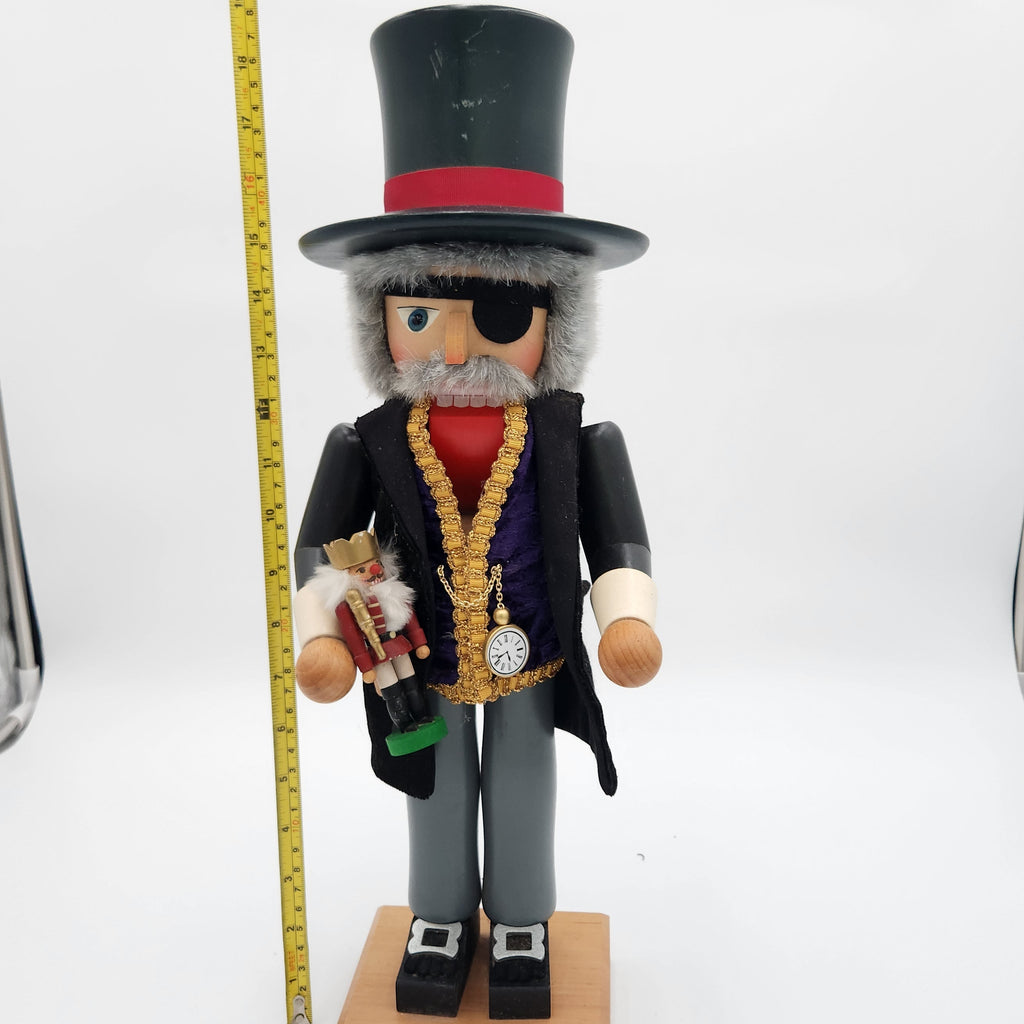 A pirate-themed wooden nutcracker figurine with a black top hat, eyepatch, gray beard, and black coat over a purple vest, holding a small toy soldier. A yellow tape measure is shown next to it for scale against a white studio background. Christian Ulbrickt