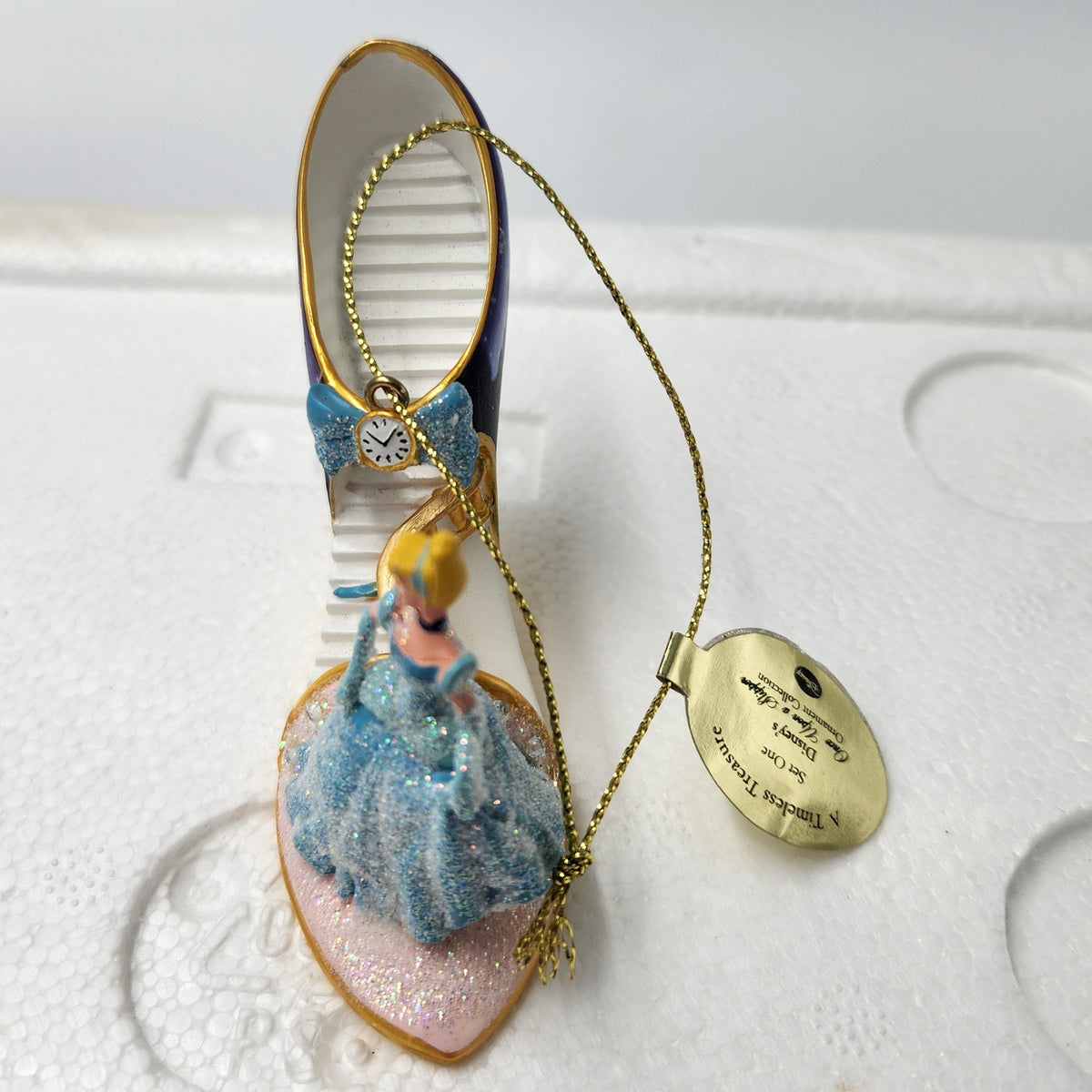 A Cinderella-themed ornament featuring a miniature Cinderella figurine in a blue gown on a shoe-shaped base, with a gold hanging chain and a round gold tag, photographed on white foam packaging.