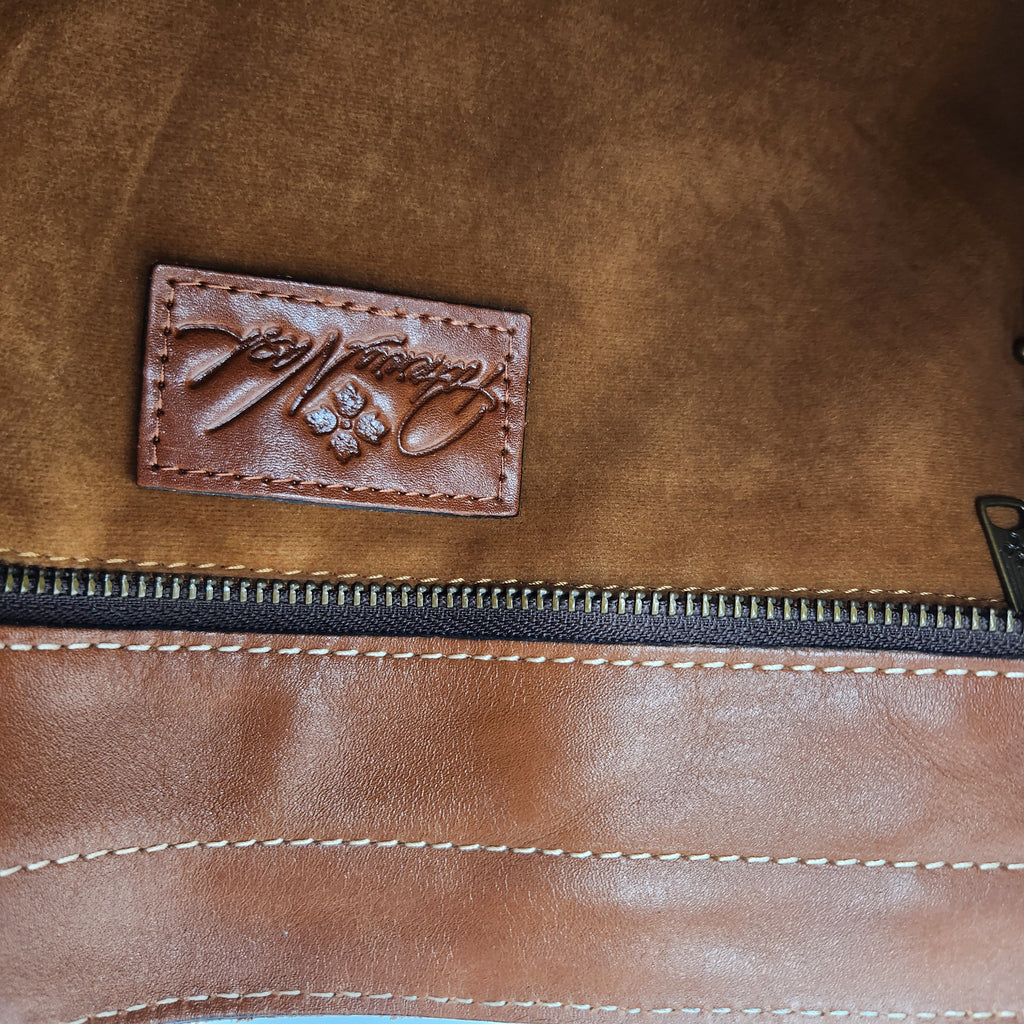 Close-up of a brown Patricia Nash leather zippered pouch or bag interior, showing a stitched rectangular brand patch with cursive script and a small flower motif near the zipper.