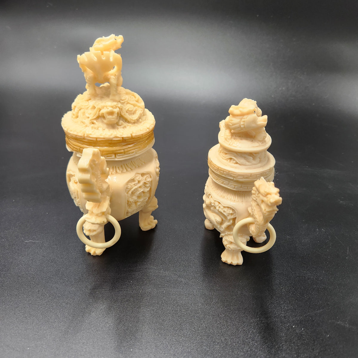 Two cream-colored carved decorative censer-like objects with lion motifs and hanging rings, displayed on a dark gray background. Vintage Chinese Carved Resin Faux Ivory Incense Burners Tripod Censers 