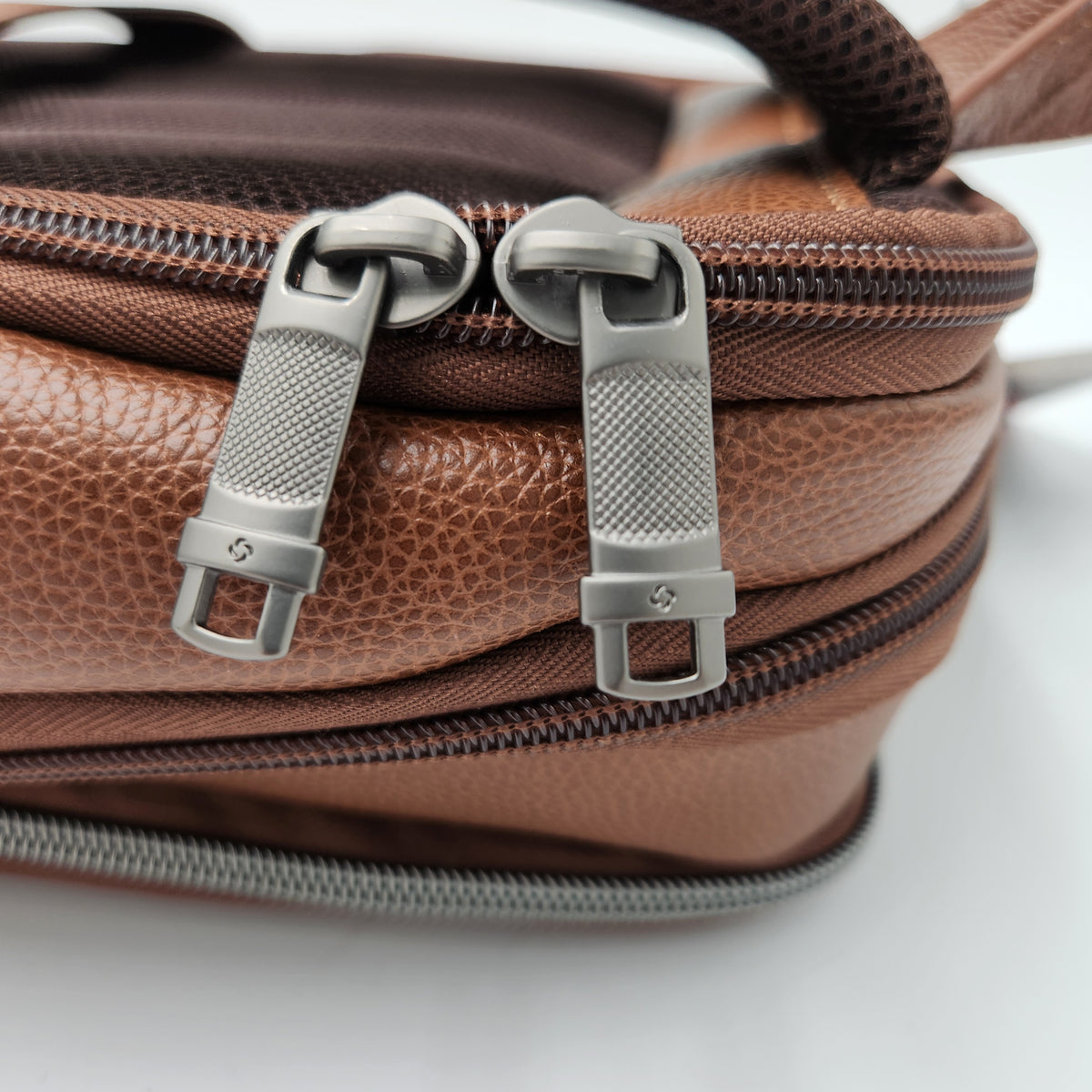Close-up of a Samsonite brown textured leather bag with two silver zipper pulls and dark brown zippers on a white background.