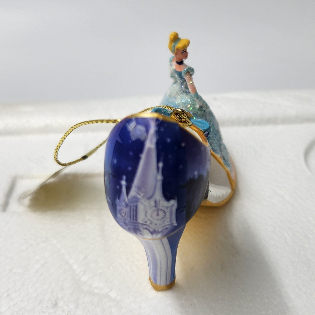 A Cinderella-themed ornament shaped like a high-heeled shoe, featuring a deep blue glittery body with a small figure of Cinderella in a light blue gown on top, attached to a gold hanging loop. The ornament is placed on a white foam packaging insert against a light gray background.