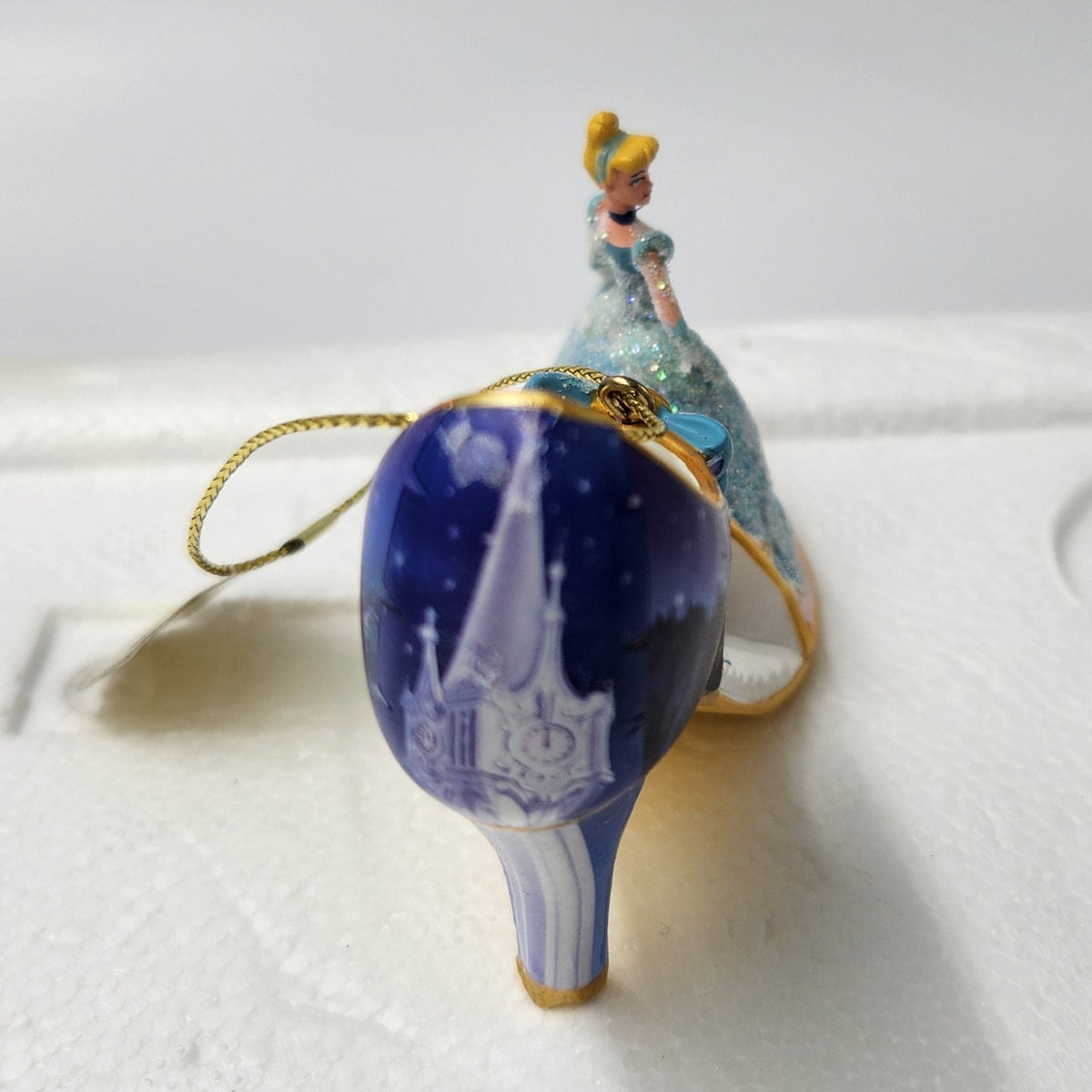 A Cinderella-themed ornament shaped like a high-heeled shoe, featuring a deep blue glittery body with a small figure of Cinderella in a light blue gown on top, attached to a gold hanging loop. The ornament is placed on a white foam packaging insert against a light gray background.